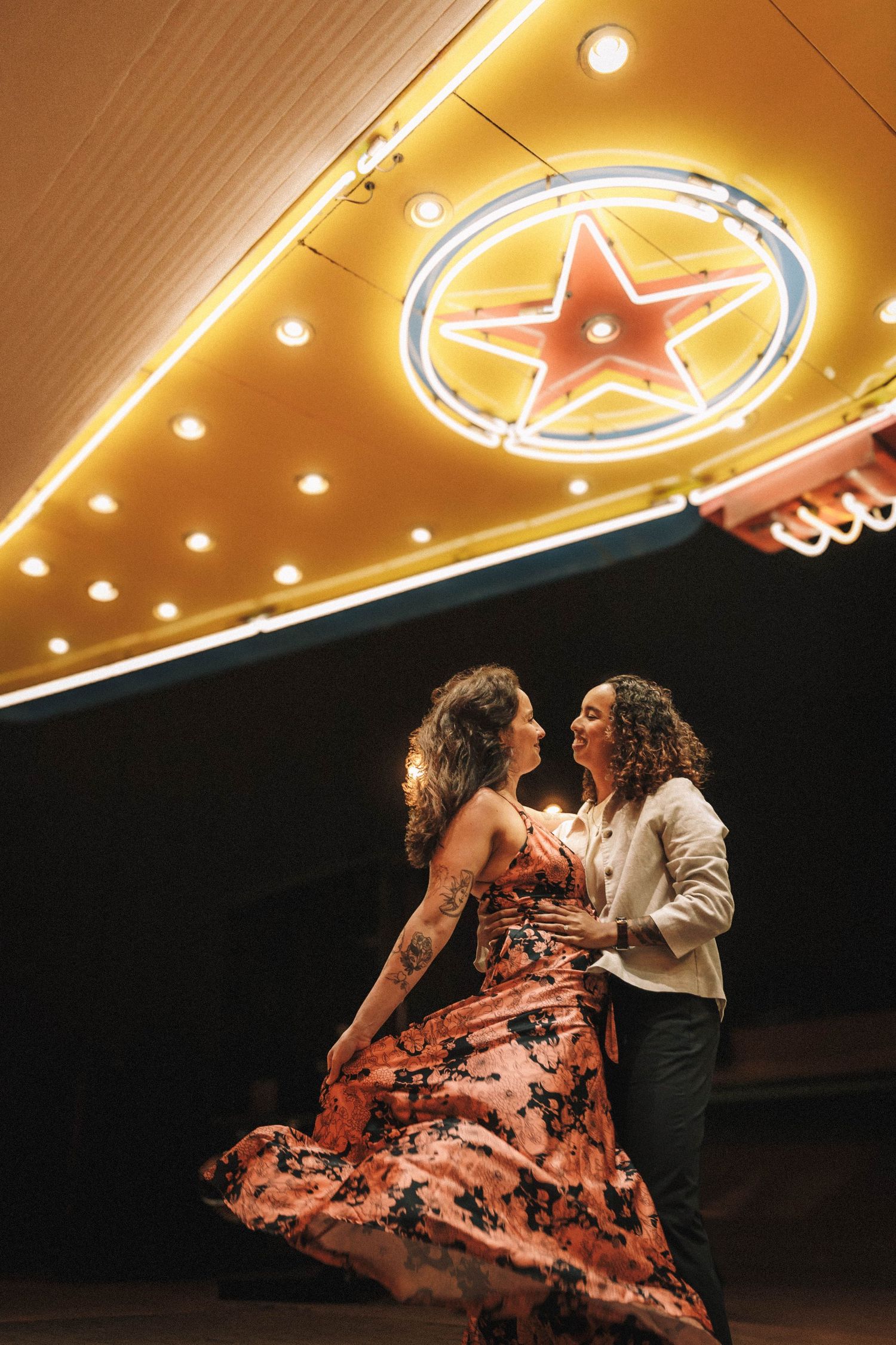 Two women dance joyfully beneath a neon star sign, one wearing a floral dress at night.