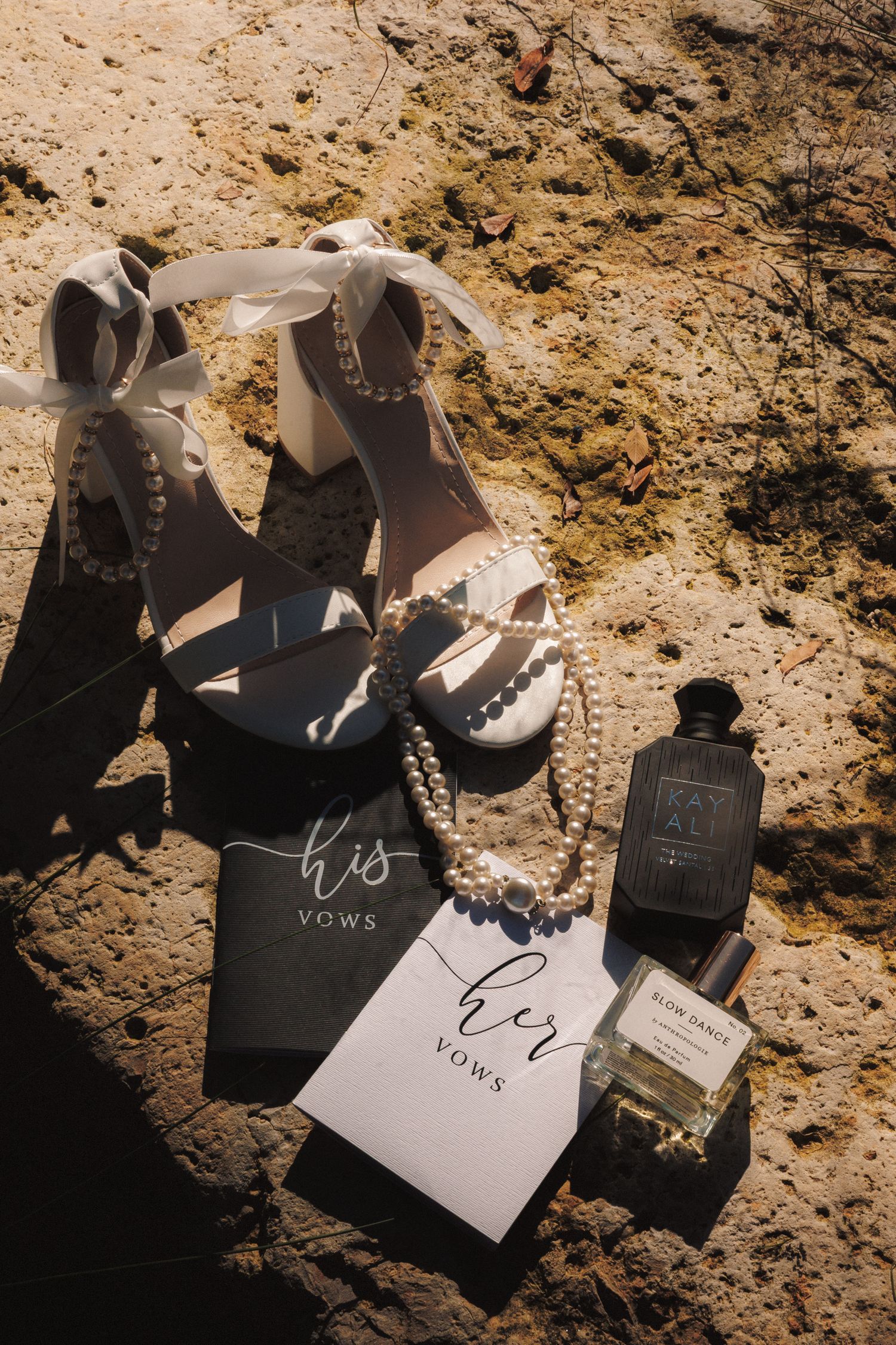 Bridal flat lay featuring white heeled sandals, pearl necklace, his and her vow books, and perfume bottles on stone.