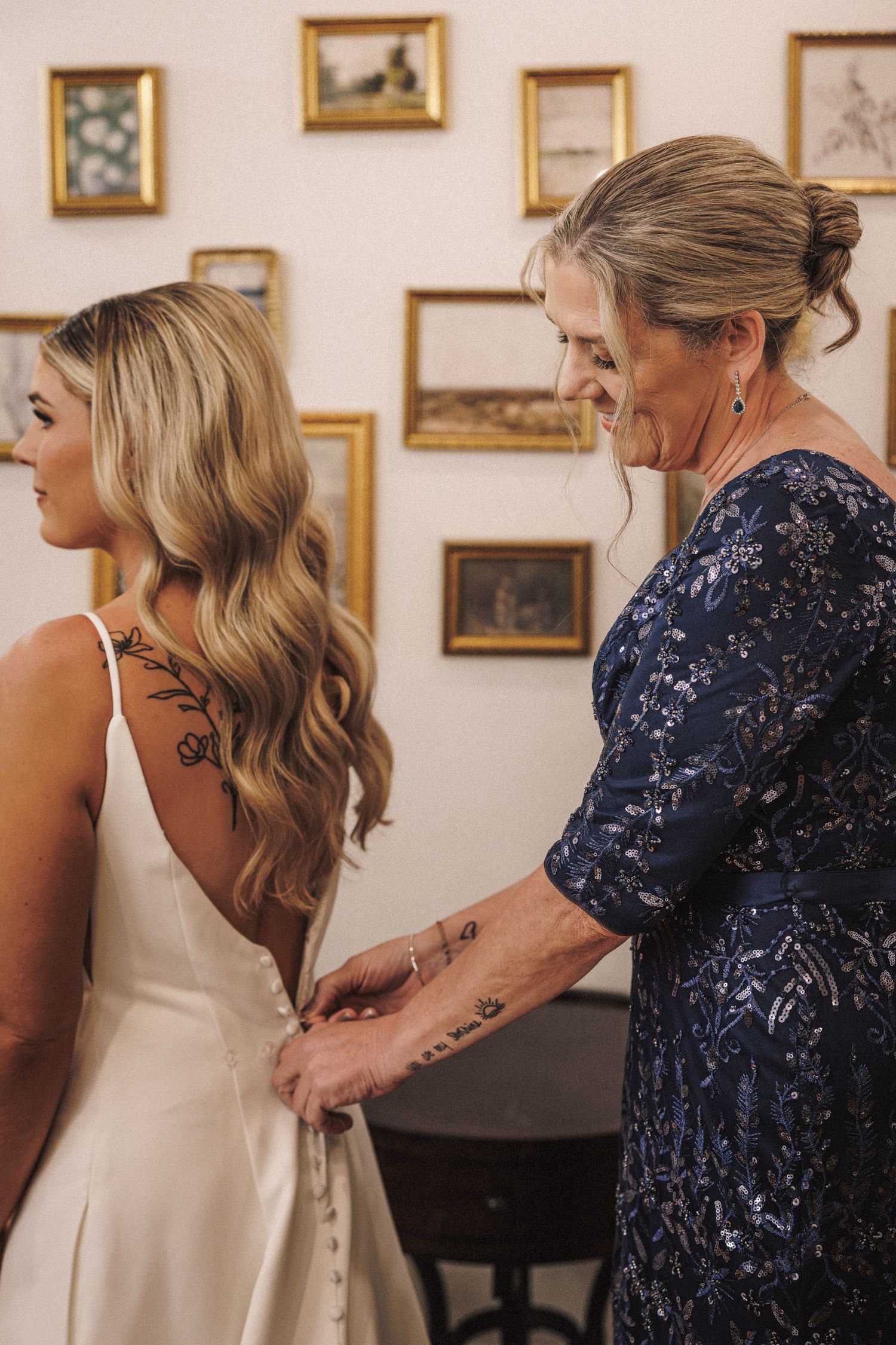 Mother in blue sequin dress carefully buttoning the back of bride's white wedding gown.
