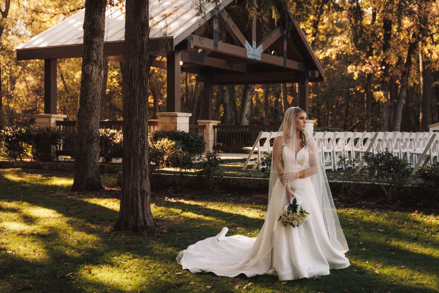 Elegant bride in white gown with veil stands on green lawn near rustic wooden pavilion in fall foliage.