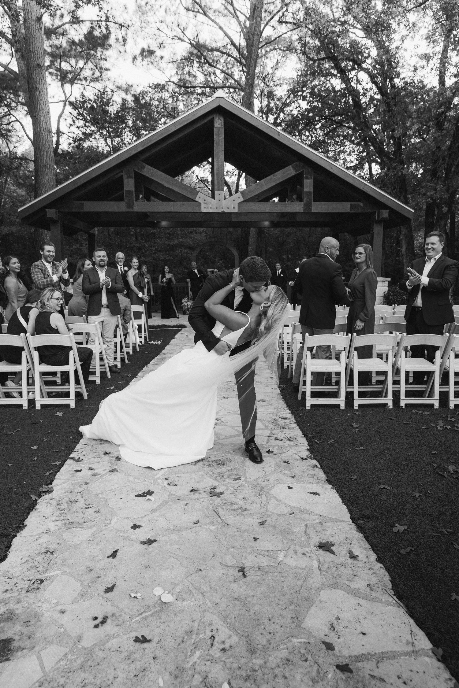 Black and white photo of groom dipping bride for a kiss in the aisle of an outdoor autumn wedding ceremony.