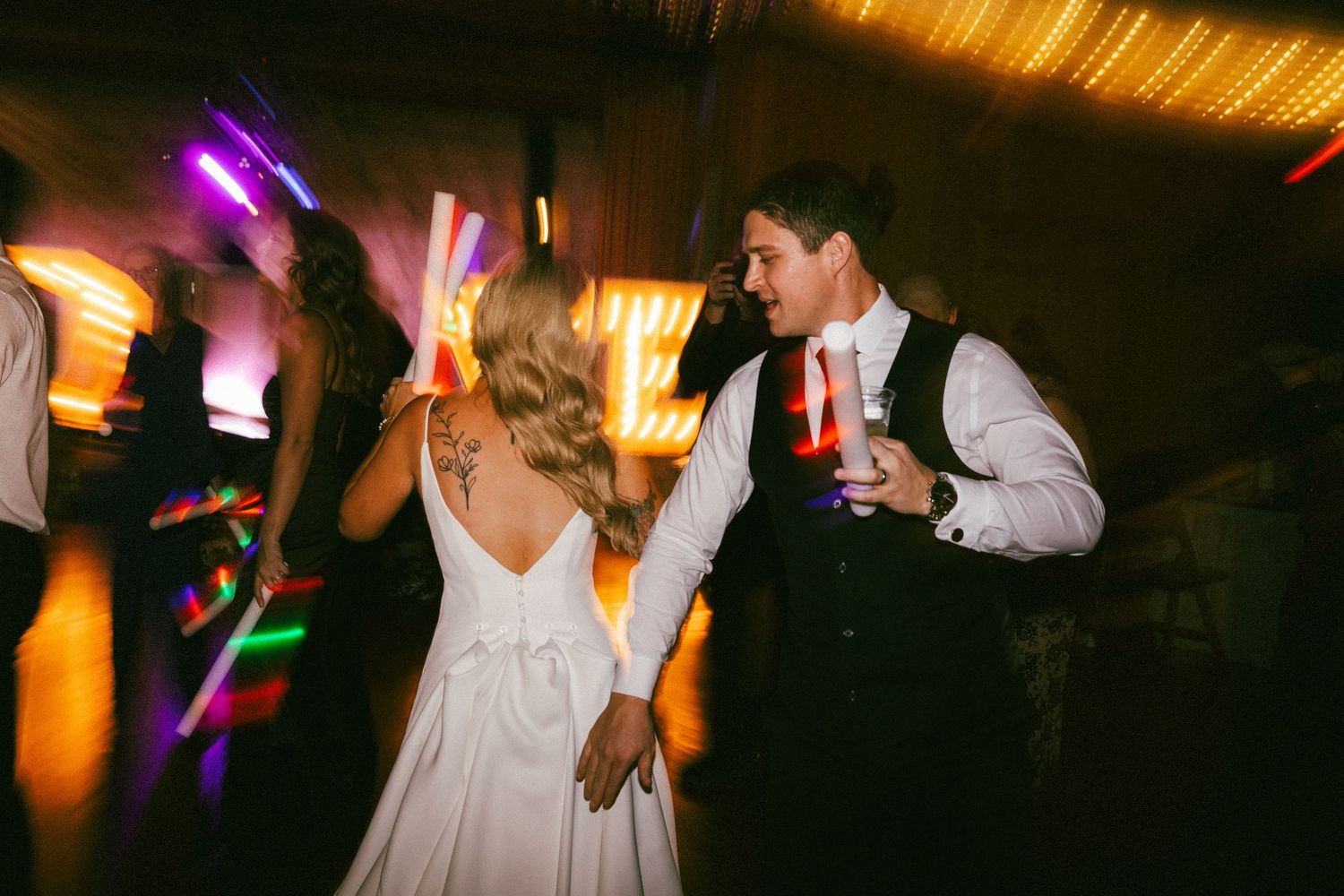 Bride and groomsman dancing together with colorful glow sticks at vibrant wedding reception party.