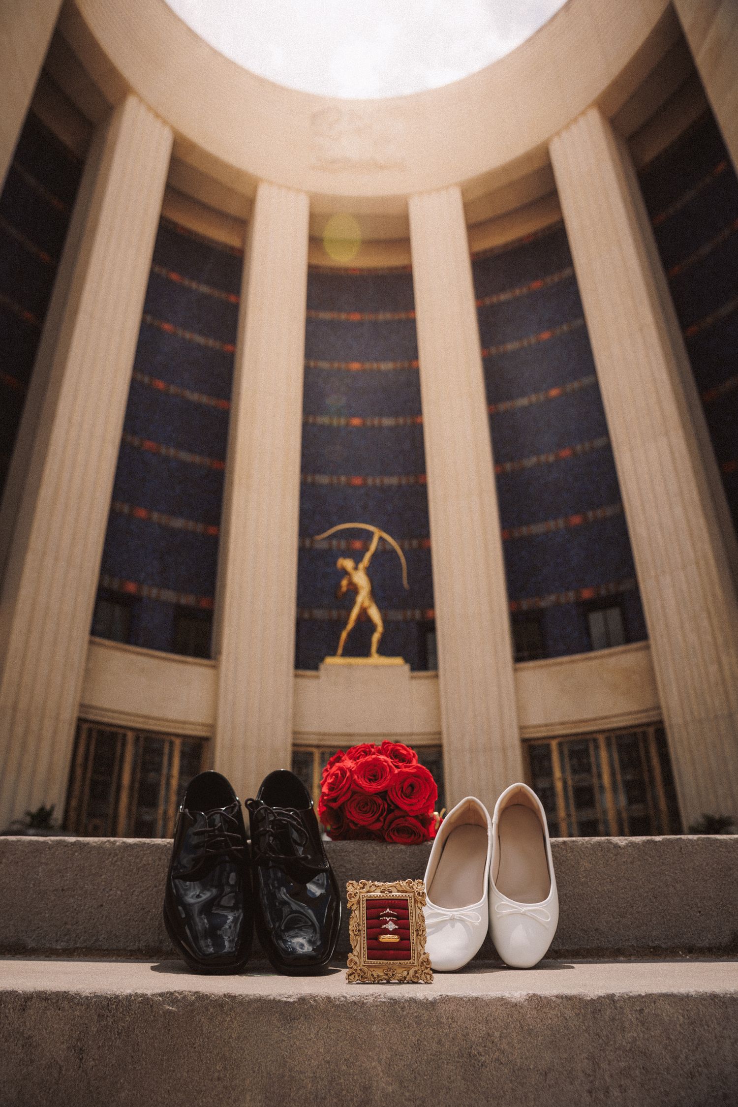 Wedding shoes, red rose bouquet, and rings displayed on steps before a grand columned building with golden statue.