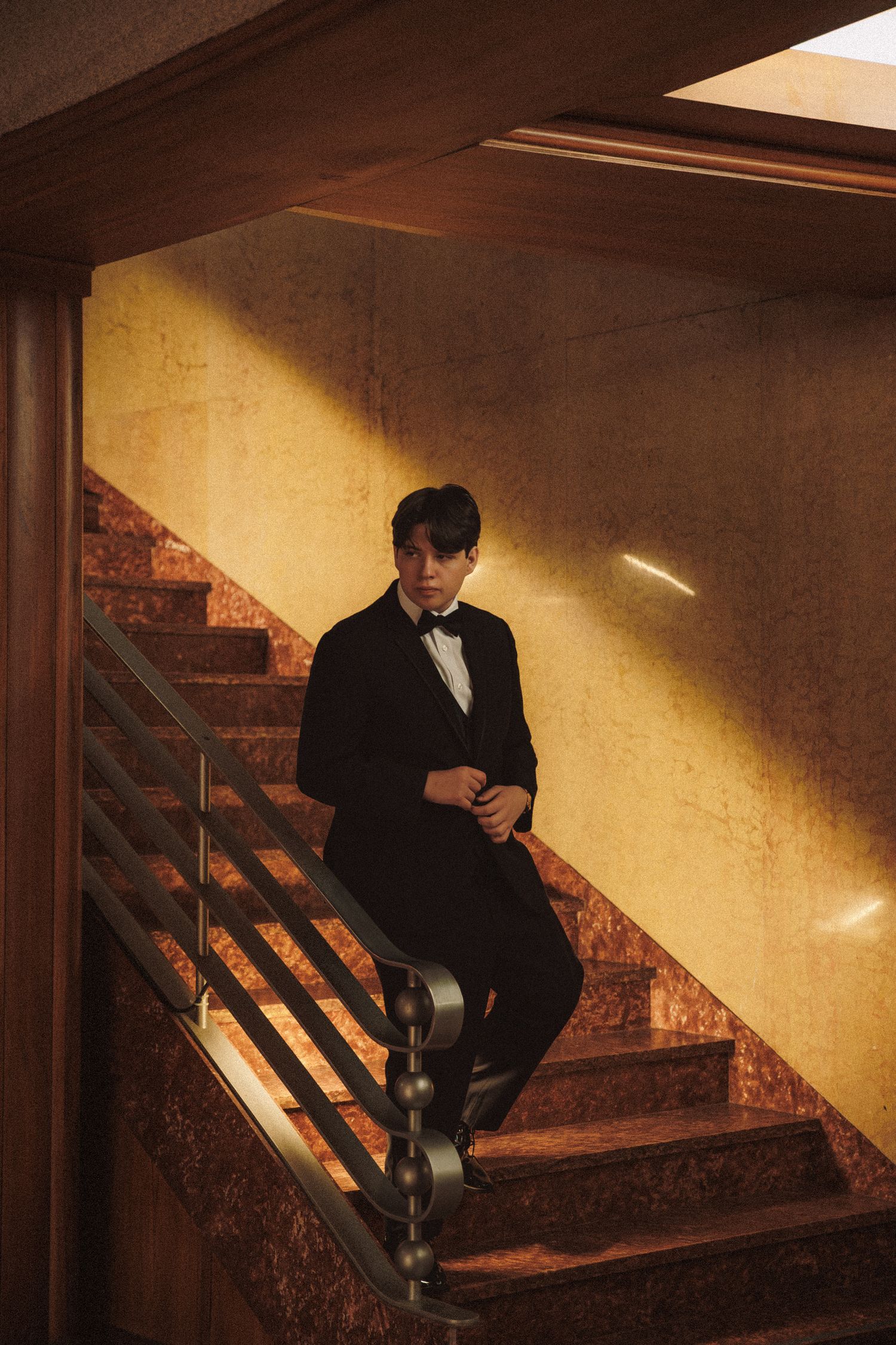 Groom in black tuxedo descends marble staircase bathed in warm golden sunlight.