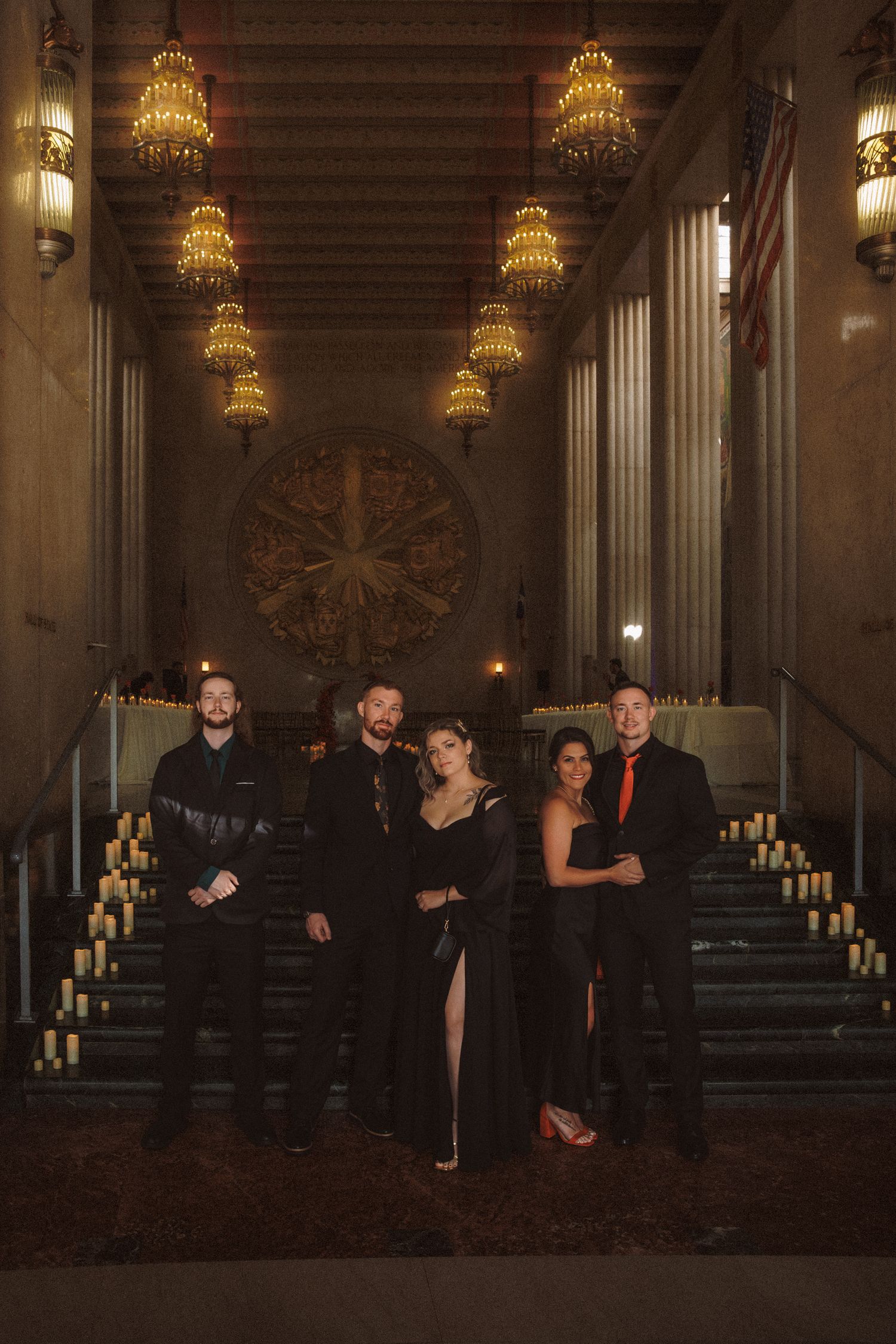 Wedding party in formal black attire posing in grand chandelier-lit hall with candles.