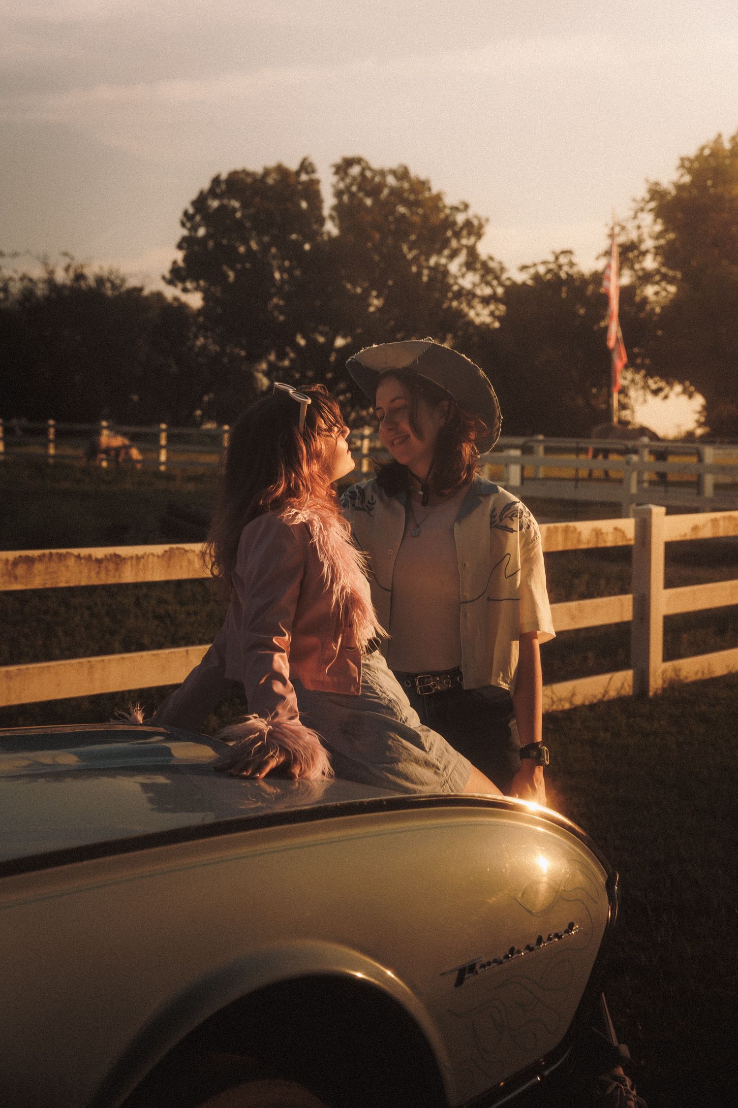 A couple sits together at sunset on a vintage convertible car parked near a white fence on a rural property.