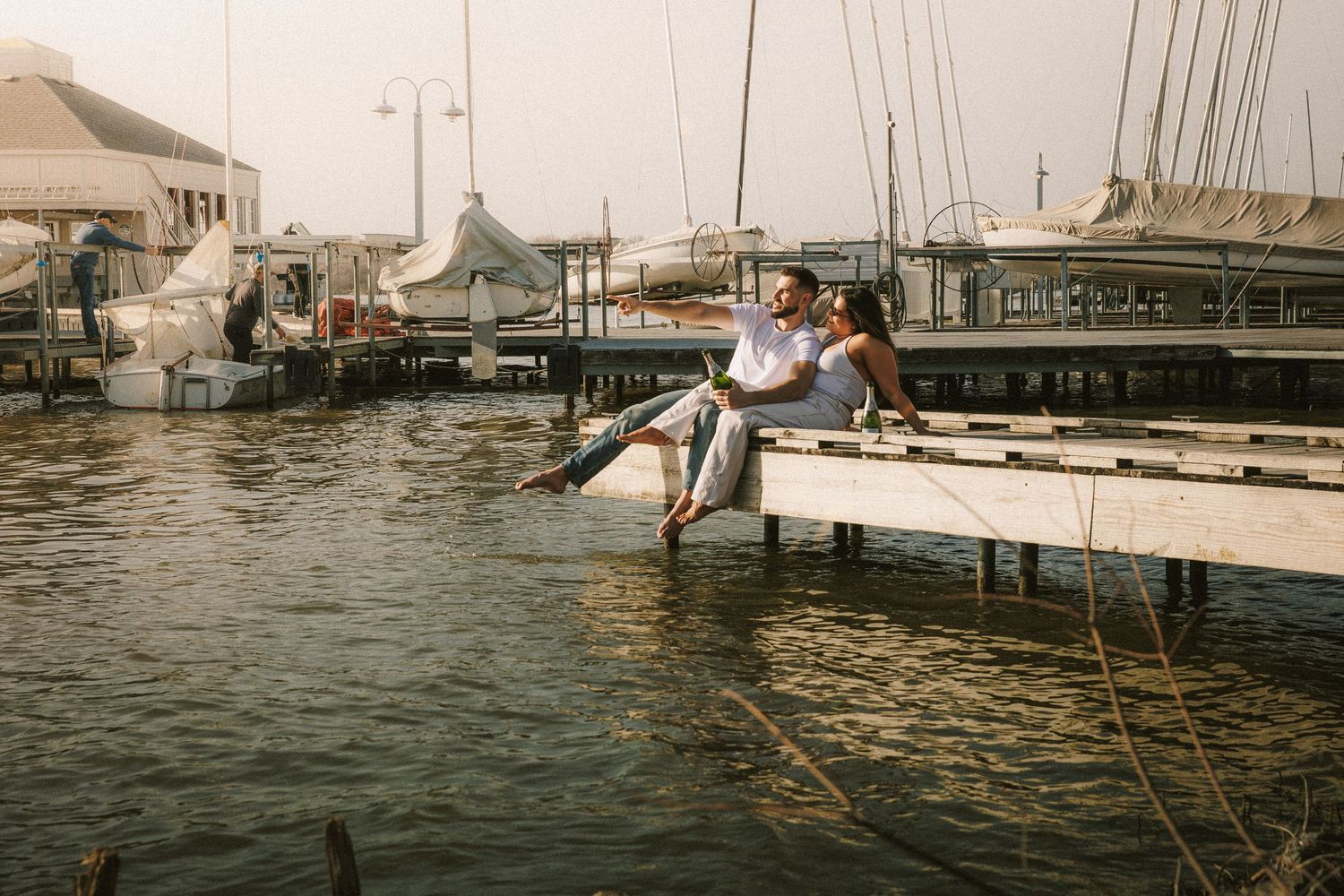 Couple sitting on wooden dock overlooking marina with boats and buildings in background.
