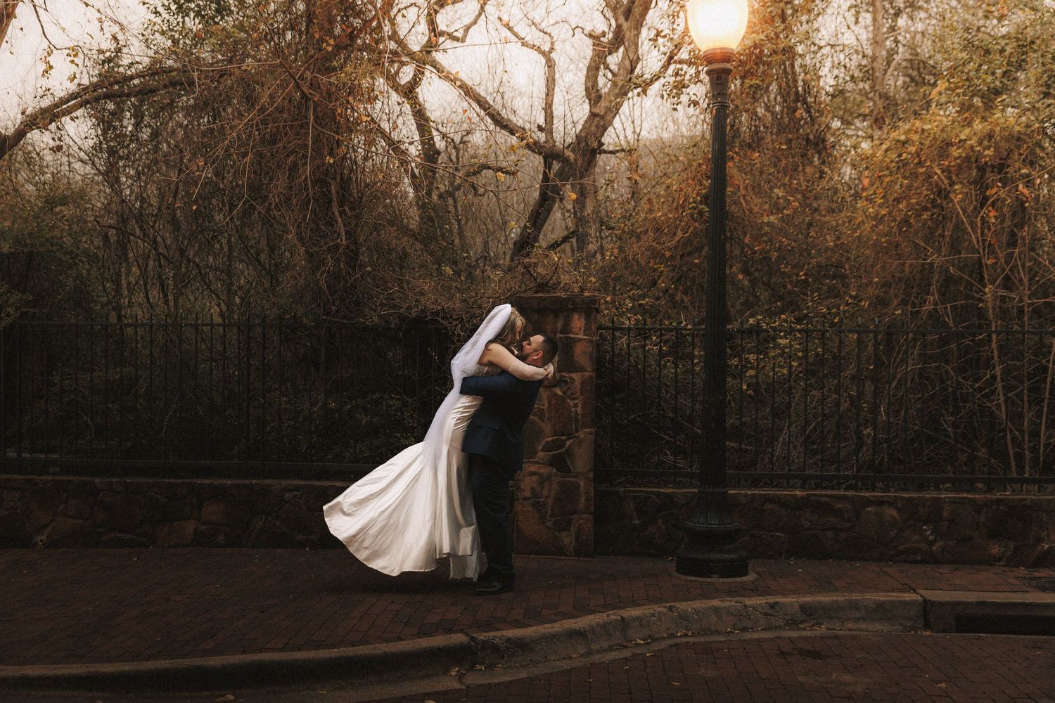 A couple in wedding attire dances in a romantic embrace at sunset in a wooded park setting.