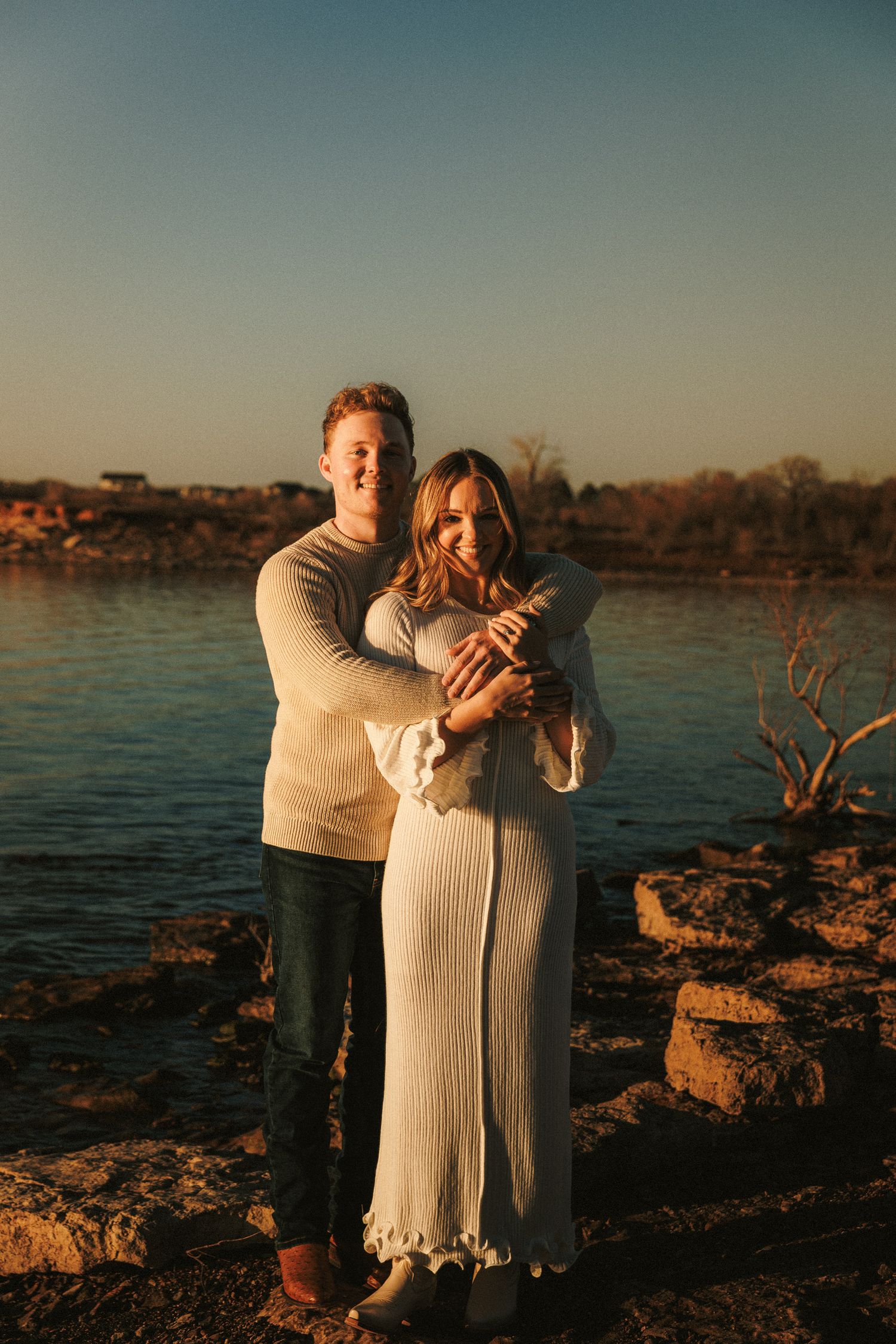 Couple embracing on rocky waterfront during sunset.
