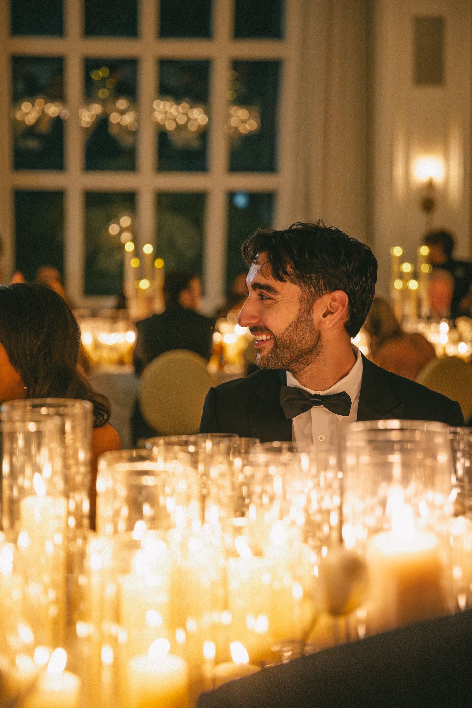 Smiling groom in black tuxedo and bow tie seated at candlelit wedding reception table with warm glow.