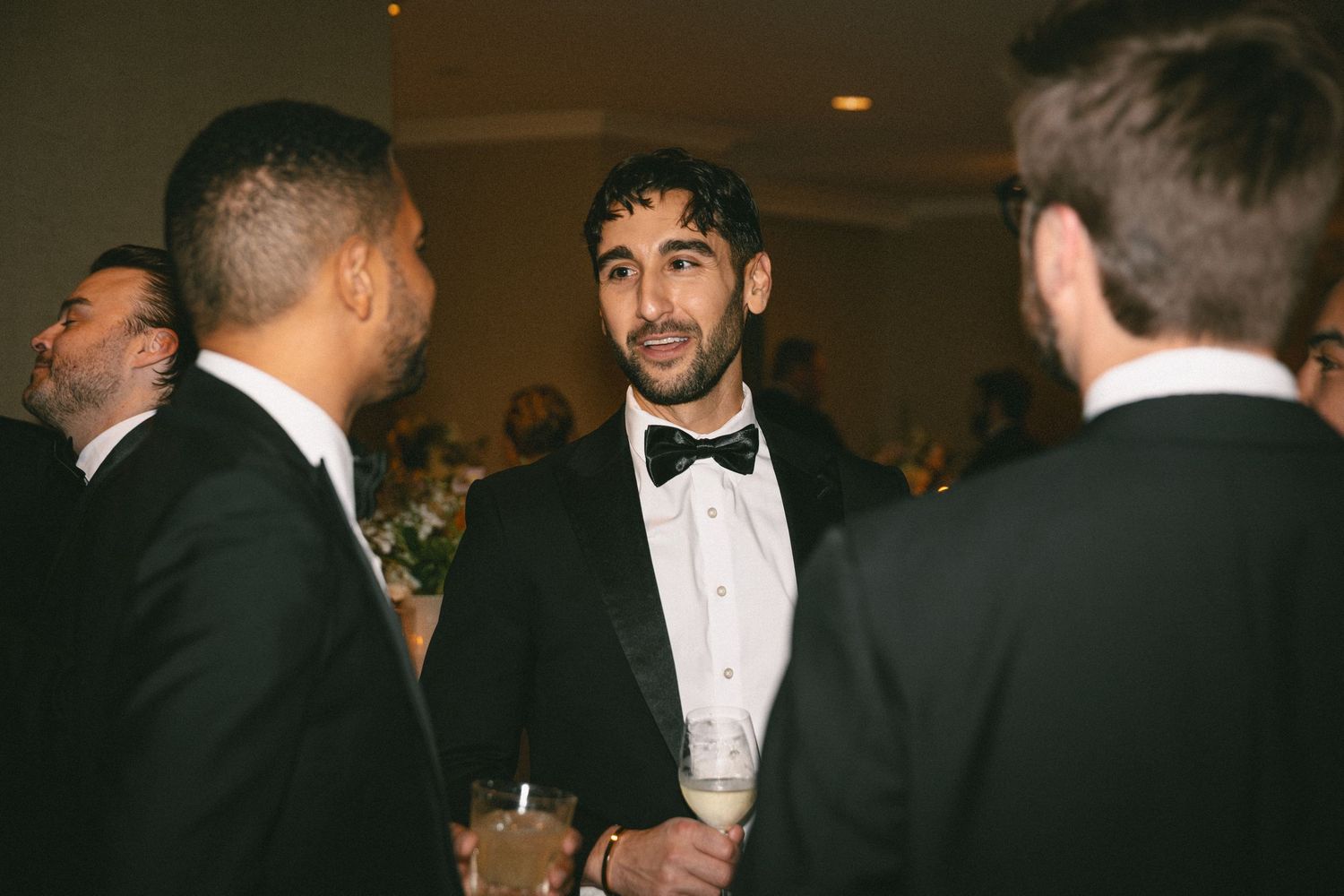 Groom in black tuxedo and bow tie socializing with guests at elegant wedding reception.