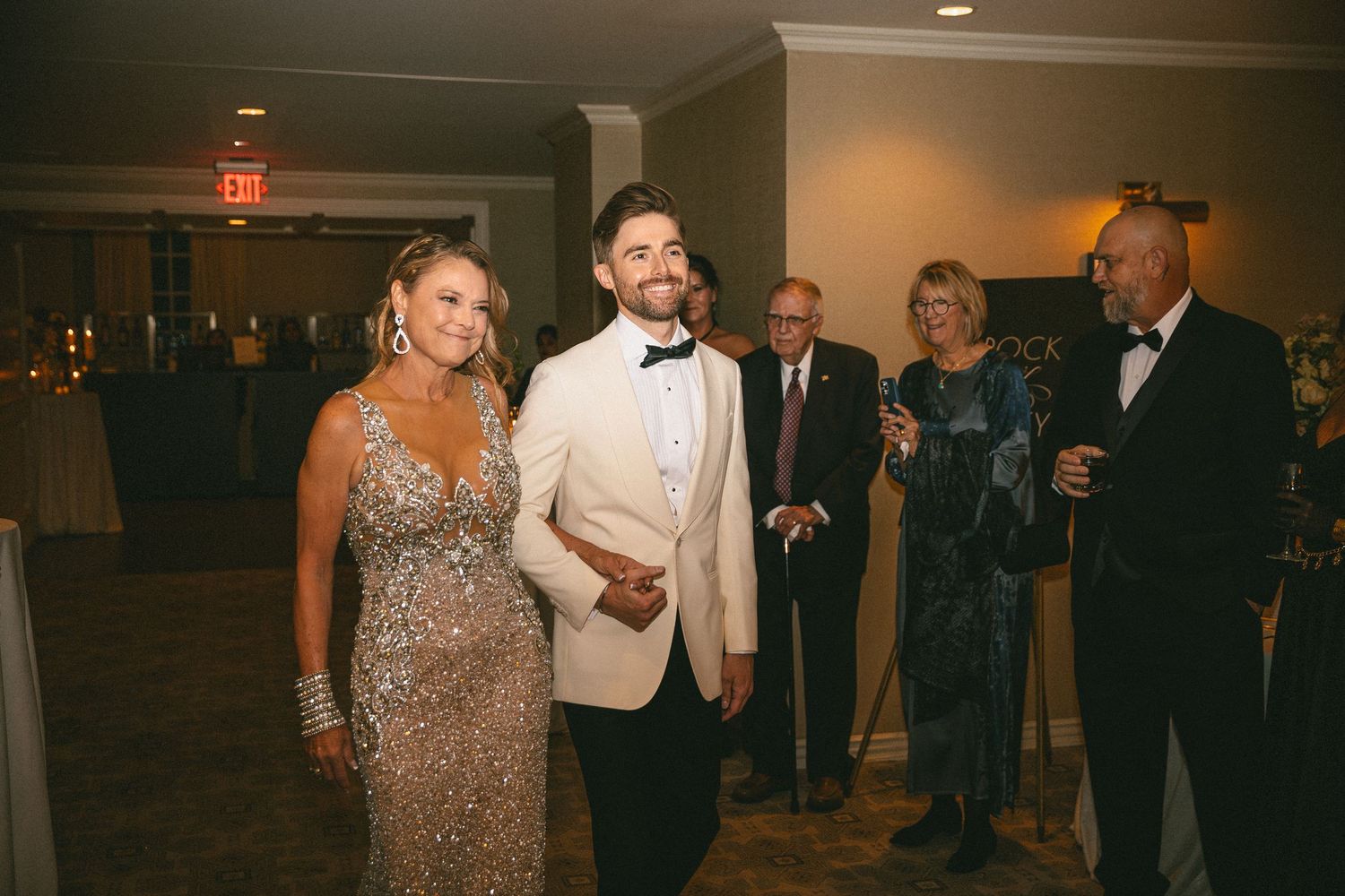 Two grooms in matching ivory tuxedos smiling as they enter their wedding reception surrounded by guests.