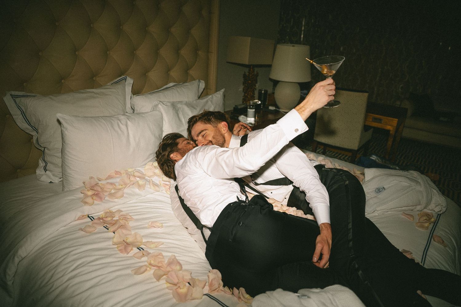 Newlywed couple relaxes on hotel bed with martini glass after wedding reception.