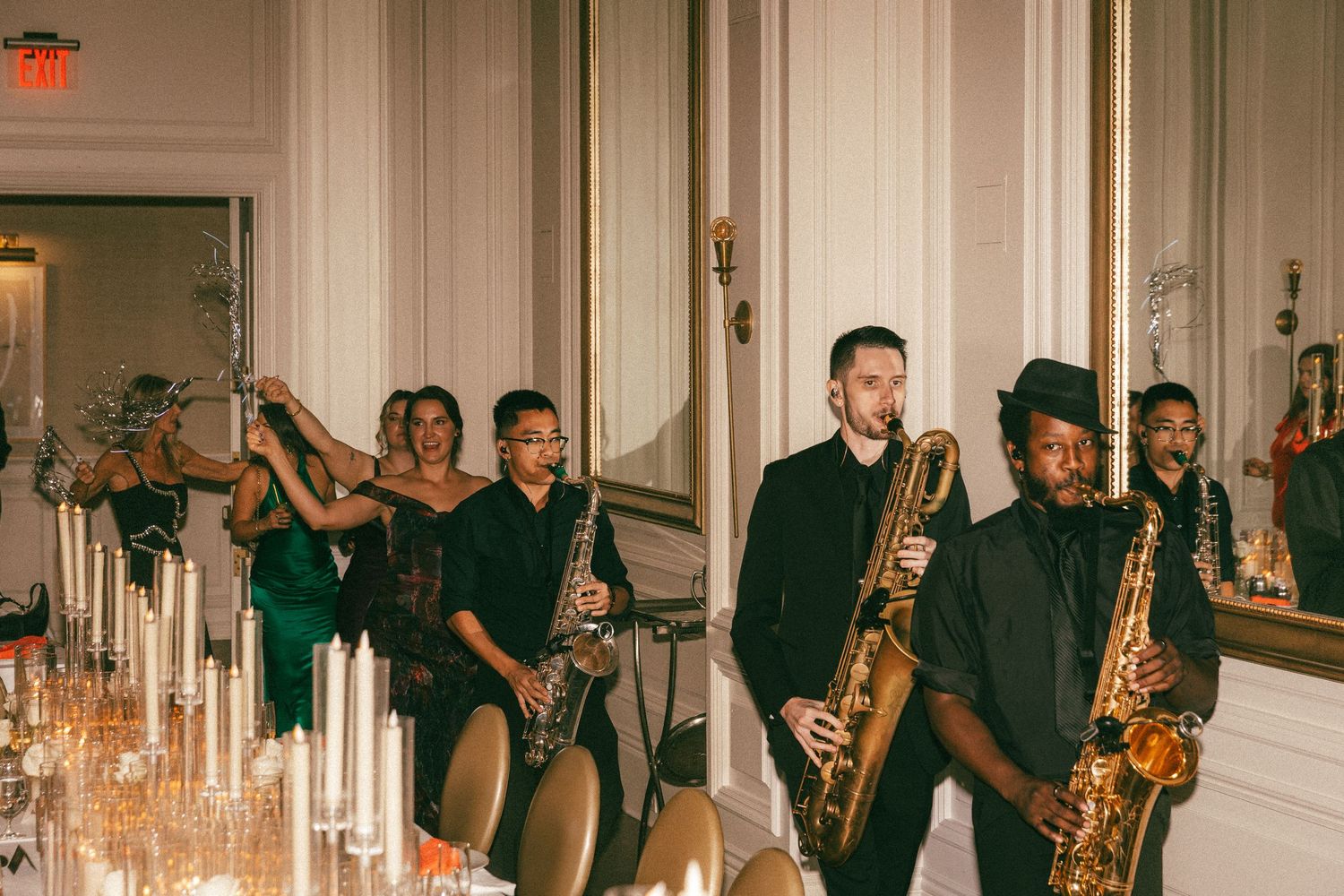 Wedding band musicians with saxophones line up beside candlelit table in ornate ballroom.