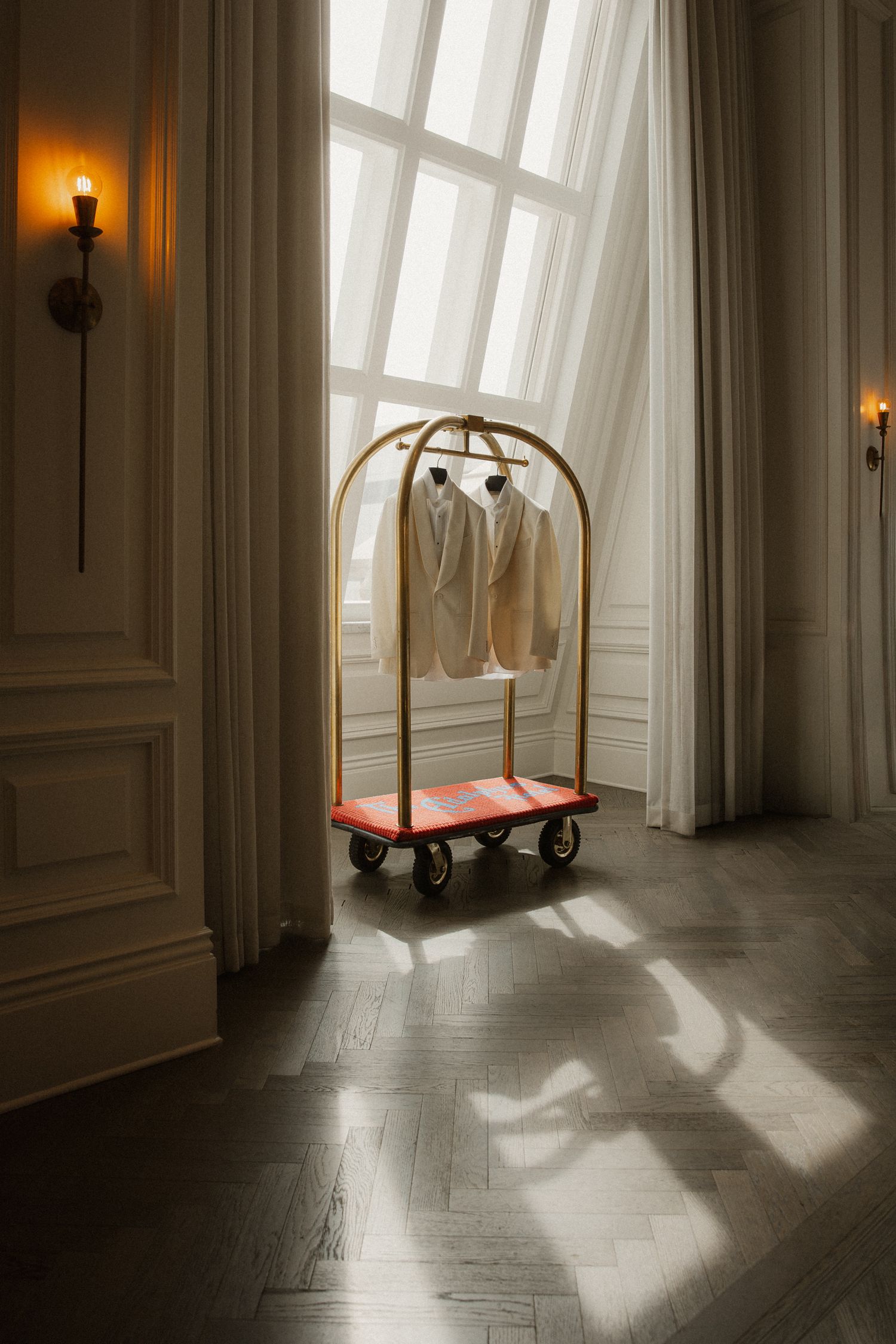 Gold hotel luggage cart with white suits hanging in sunlit elegant room with herringbone floors and wall sconces.