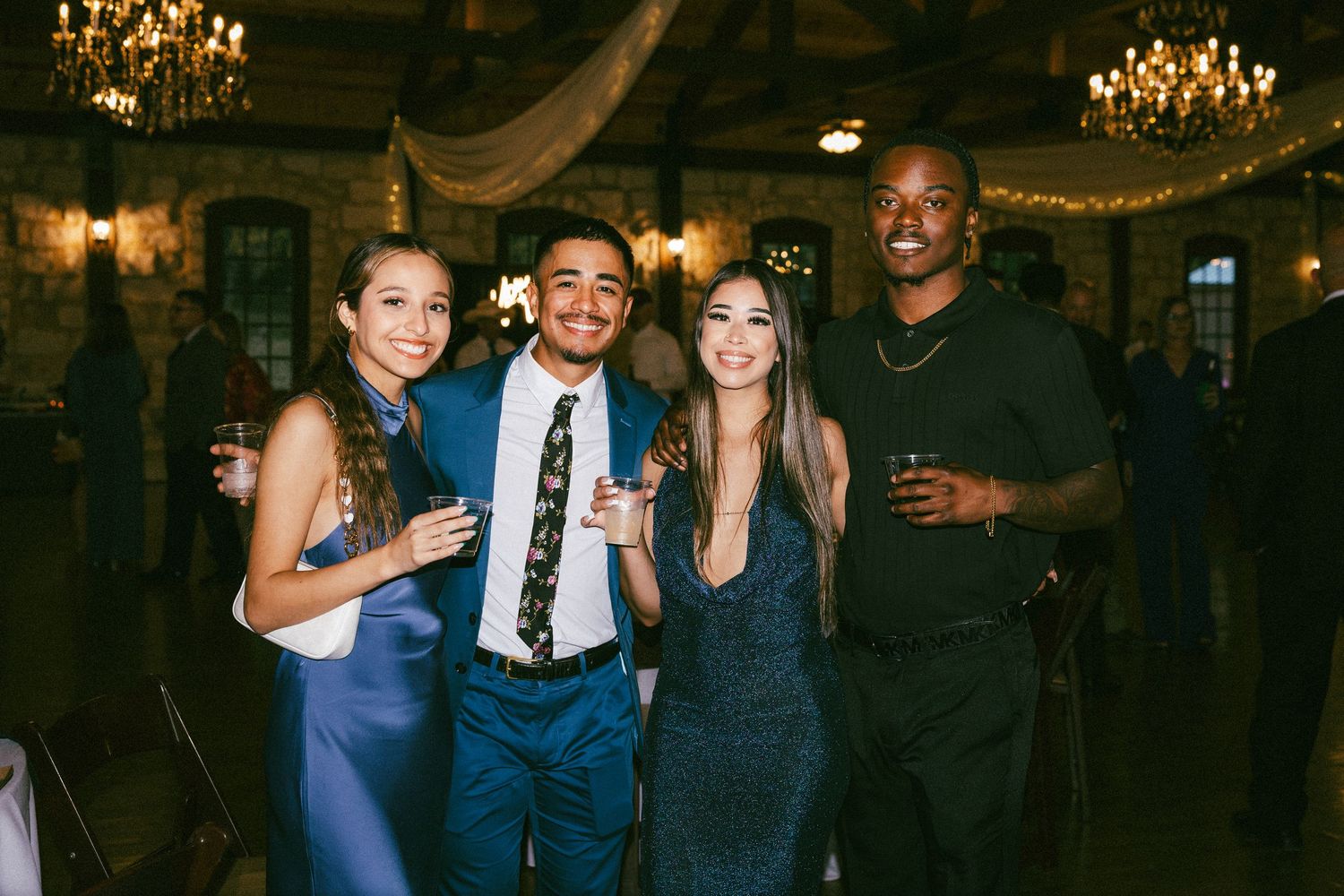 Four wedding guests smile and pose together inside elegantly lit rustic wedding reception venue.