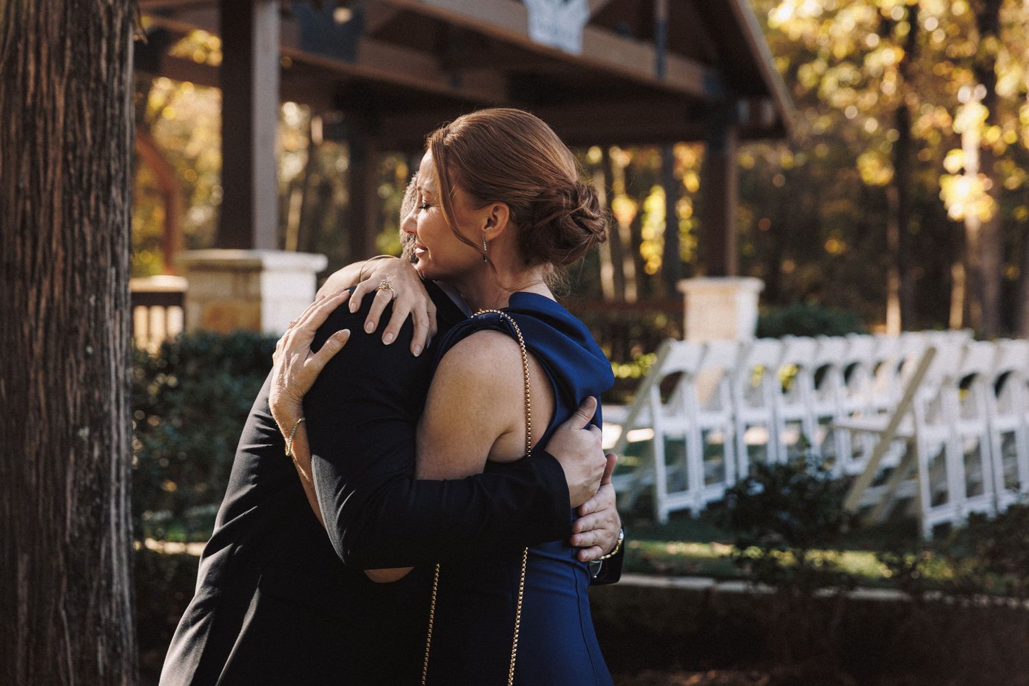 Two women sharing a heartfelt embrace outdoors near a wooded wedding ceremony venue.