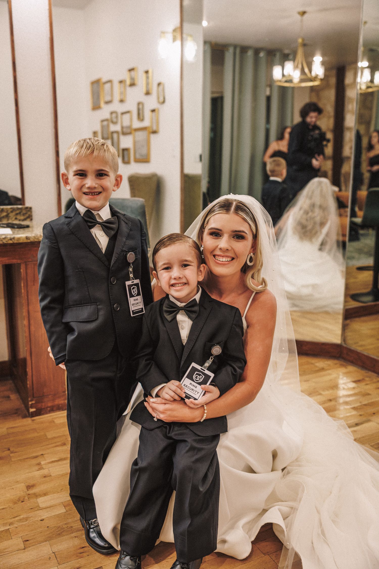 Smiling bride in white gown and veil posing with two young boys in black tuxedos at a wedding venue.