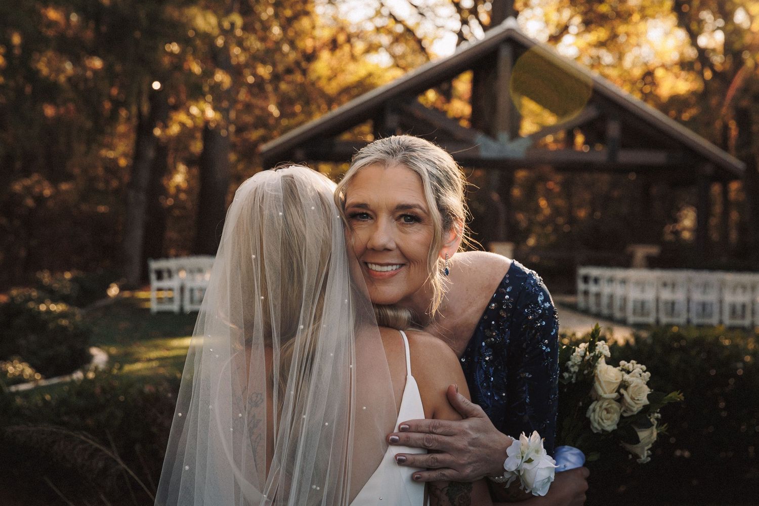 Guest embracing smiling blonde bride wearing veil at golden hour outdoor wedding ceremony.