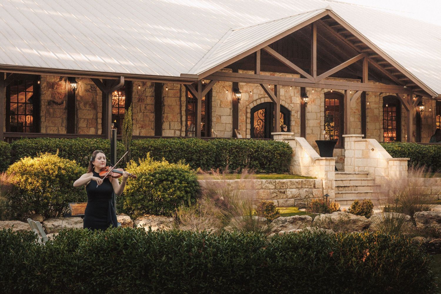 A violinist performs outdoors at a stone wedding venue surrounded by manicured hedges at dusk.
