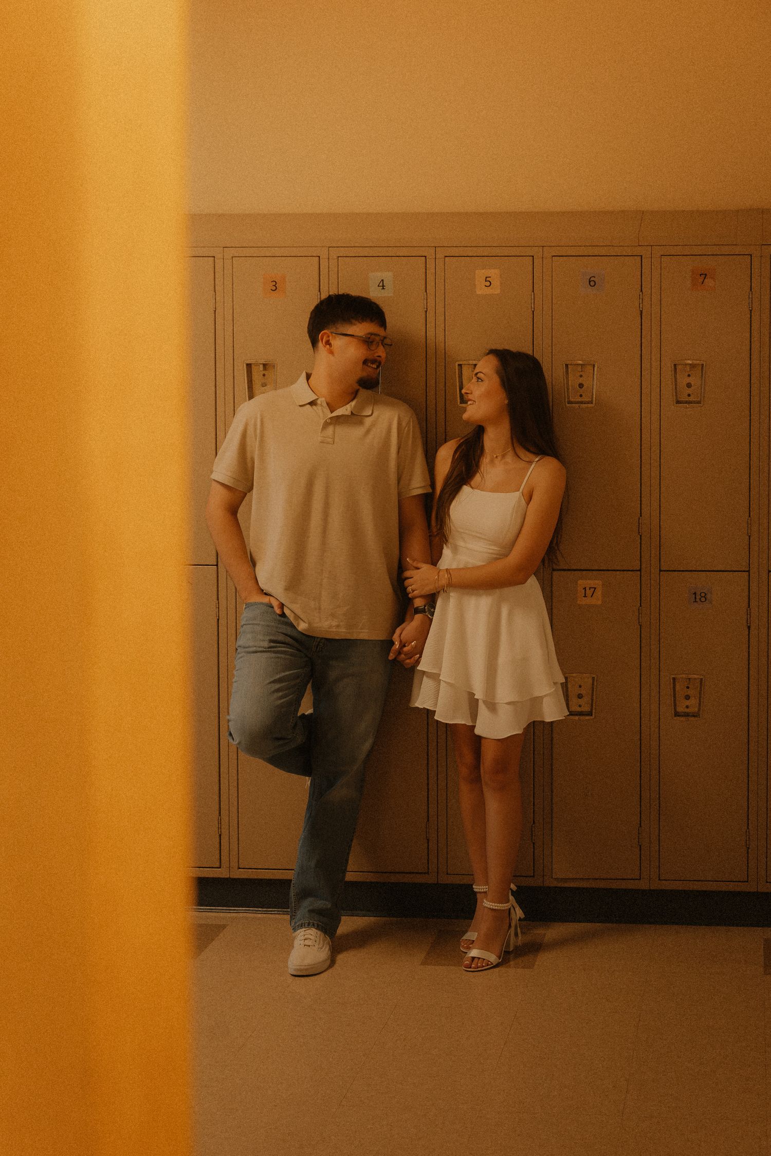 A couple holds hands and smiles at each other while leaning against school lockers in a warmly lit hallway.