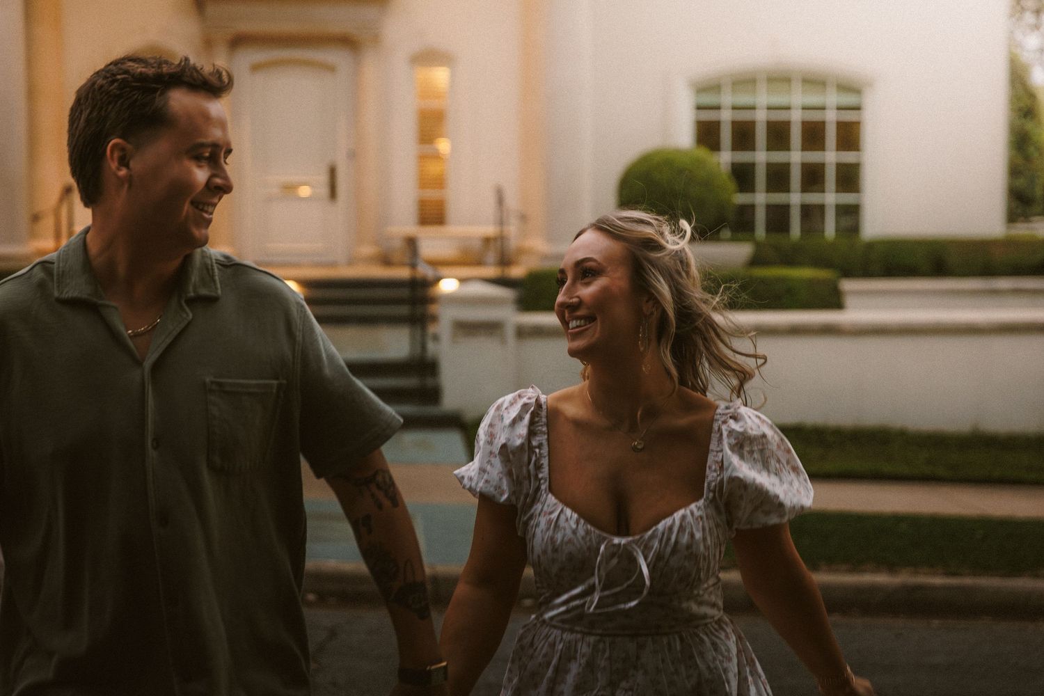 Happy couple holds hands and smiles while walking outside an elegant building at dusk.