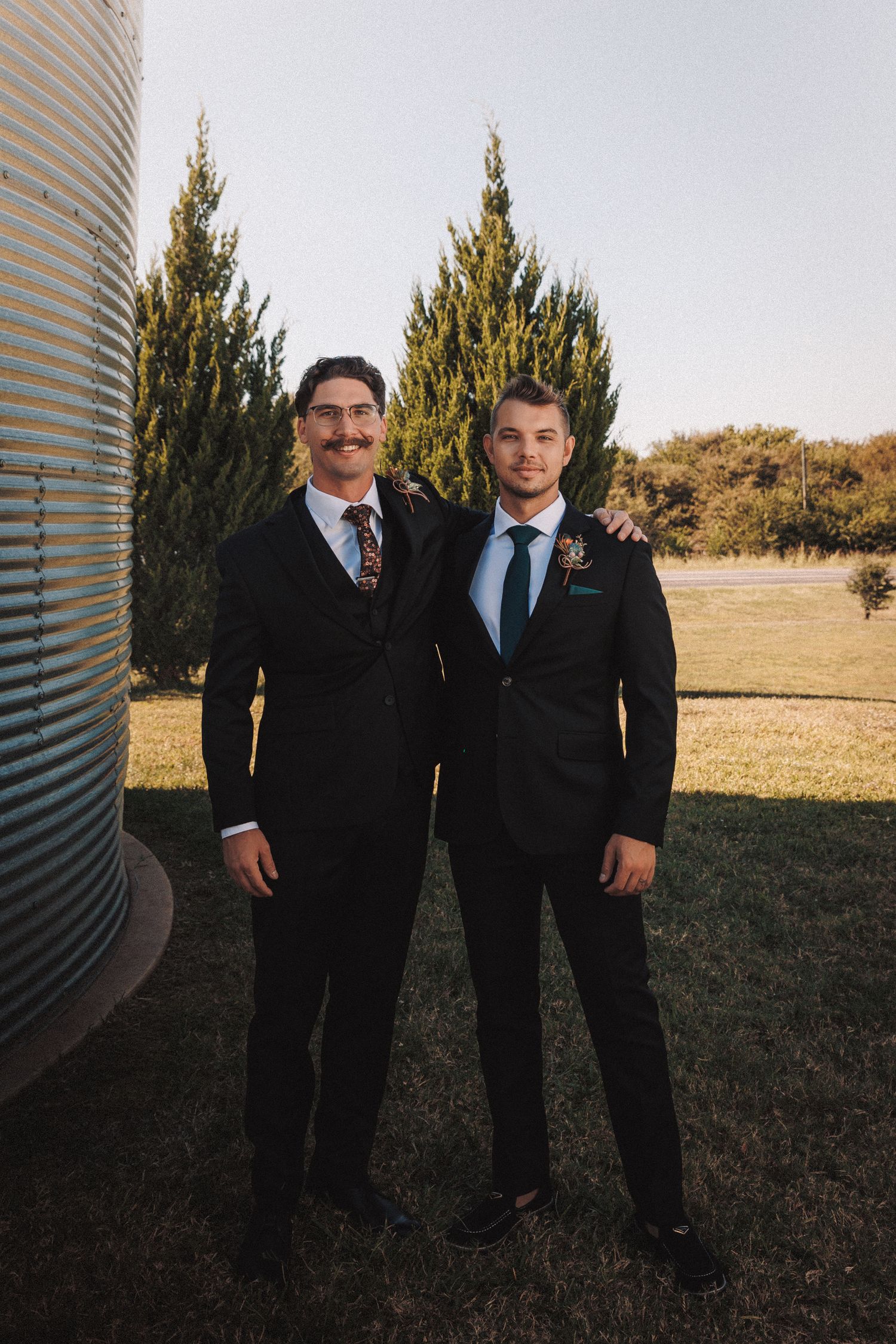 Two groomsmen in black suits posing outdoors near grain silos and evergreen trees on a wedding day.