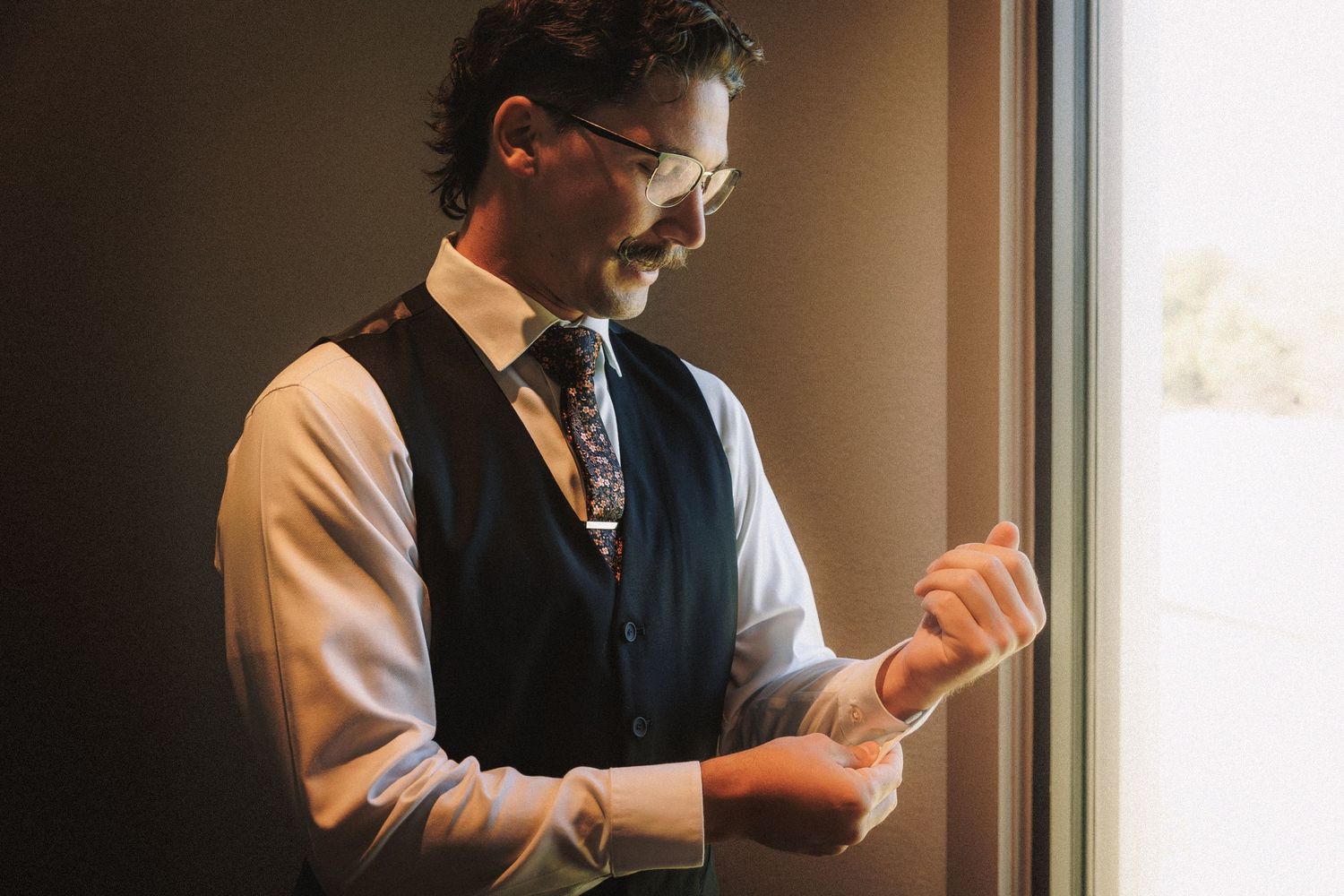 Groom in navy vest and floral tie adjusting cufflinks by window before wedding.
