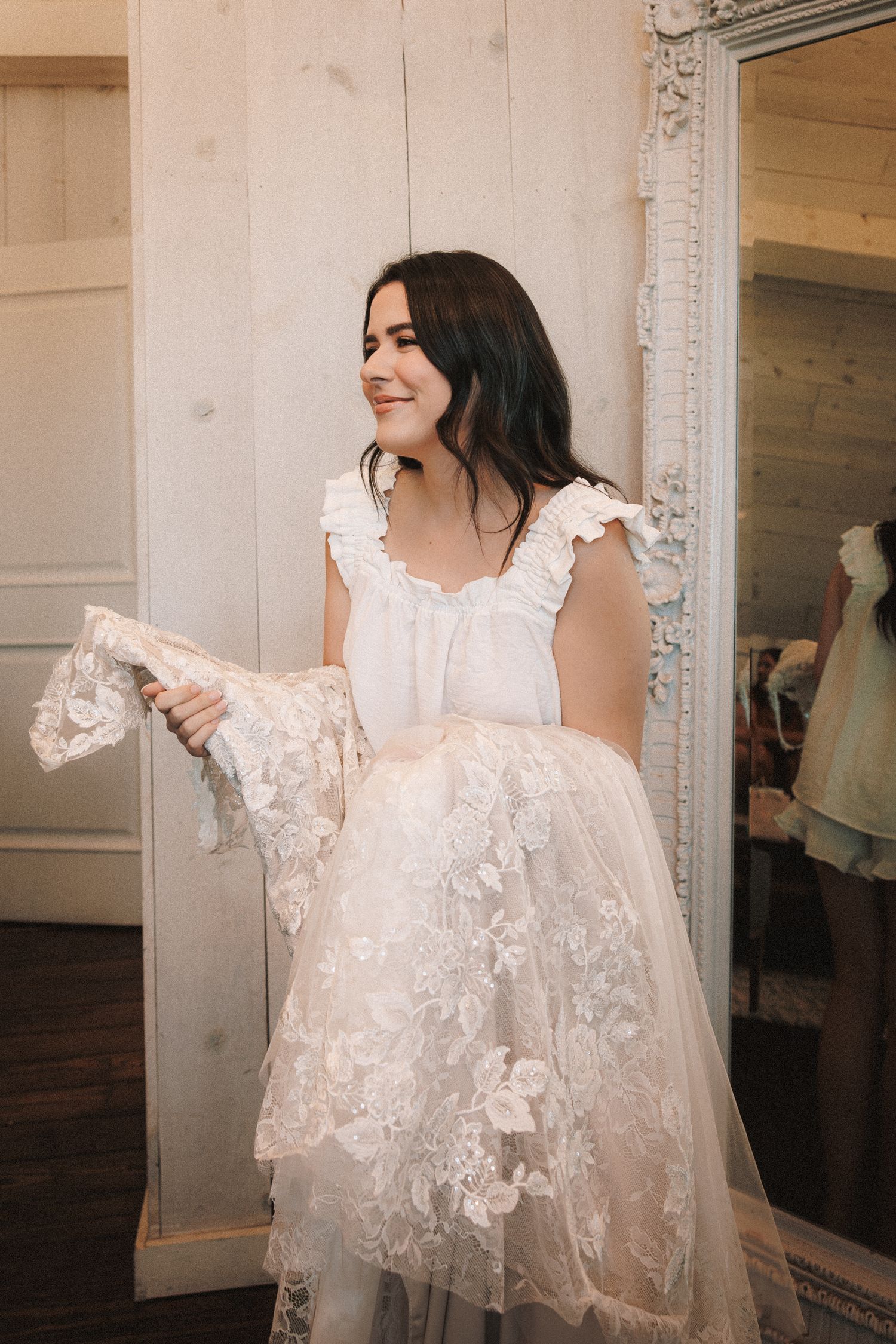 Smiling bride holding her lace wedding dress beside an ornate white mirror.