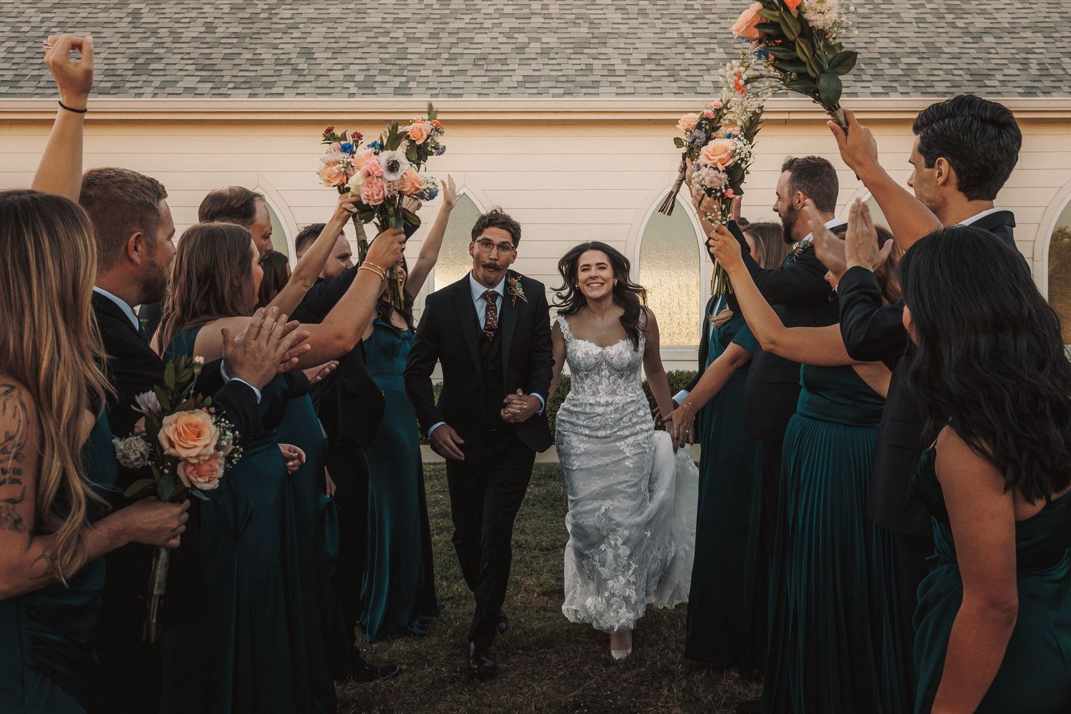 Bride and groom walk hand-in-hand through a tunnel of cheering wedding party holding floral bouquets.