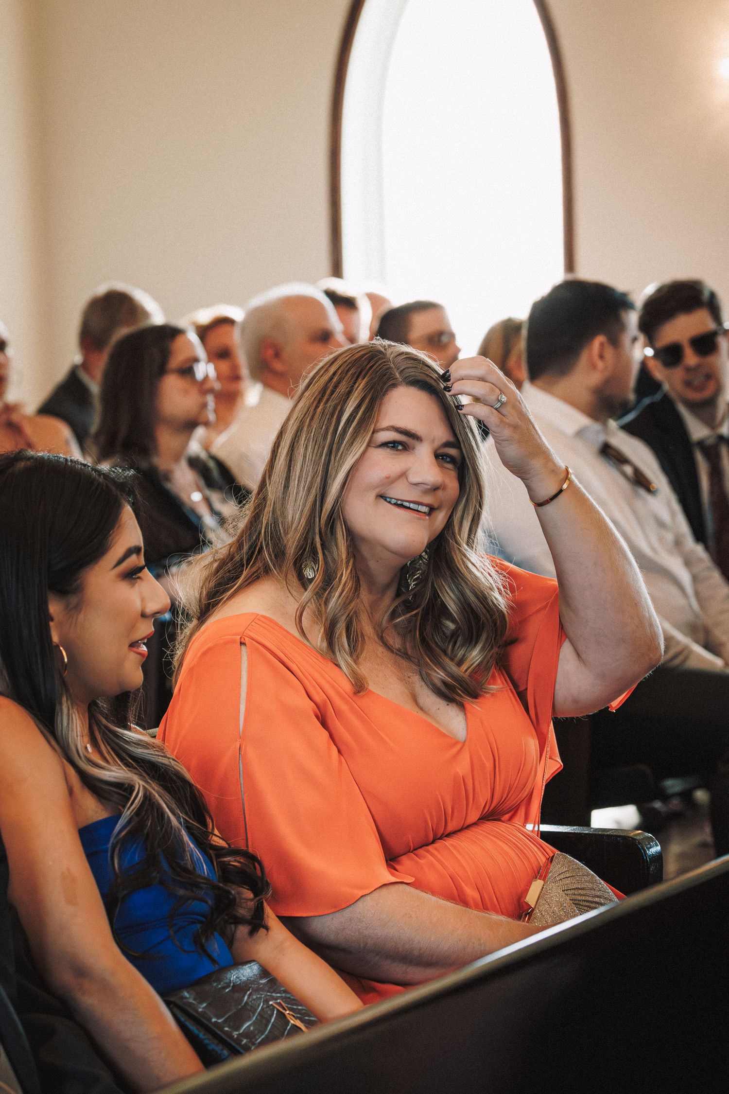 Smiling wedding guest in orange dress seated in church pew during ceremony.