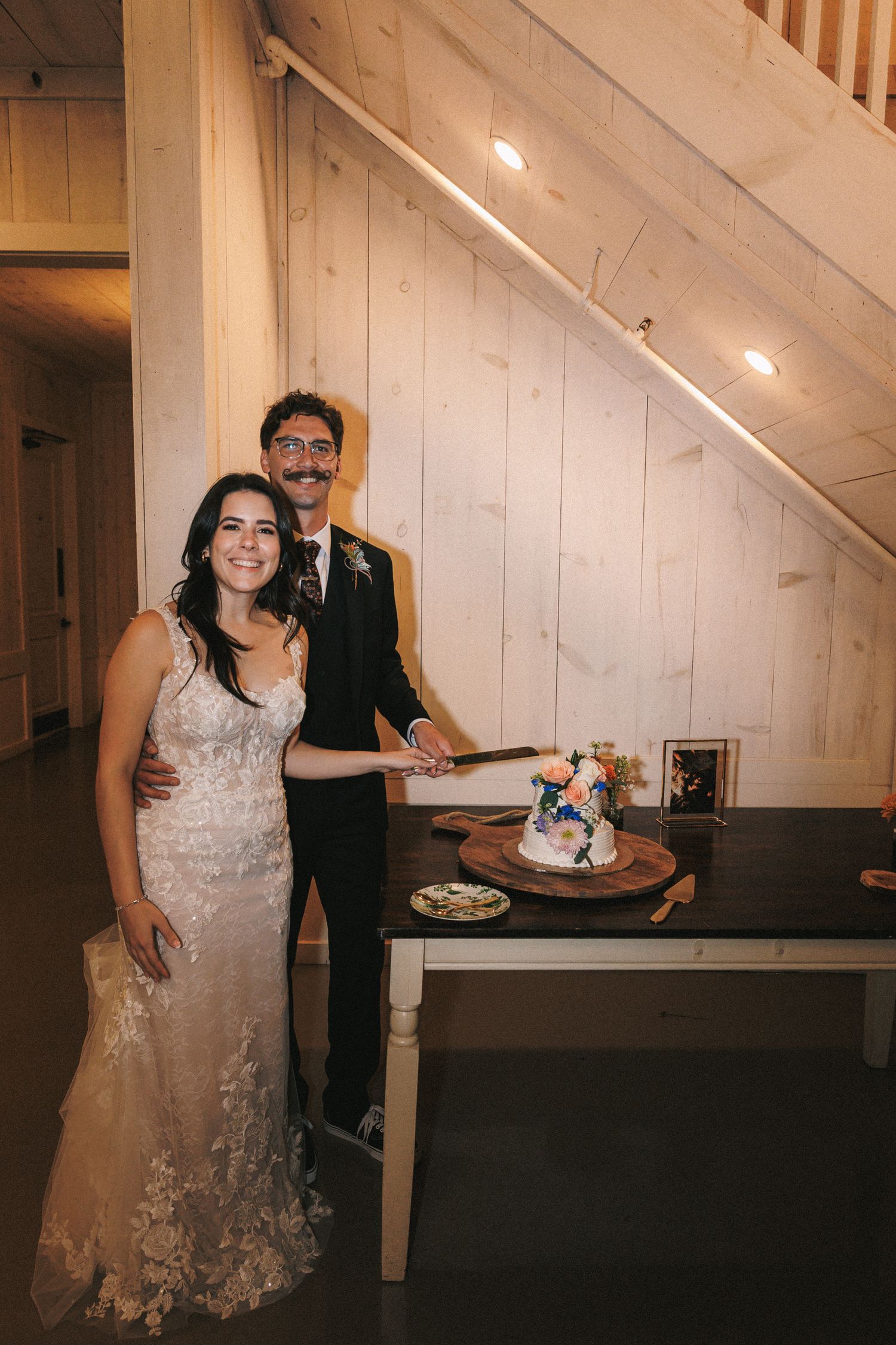 Smiling bride and groom with mustache pose together while cutting their floral wedding cake at the reception.