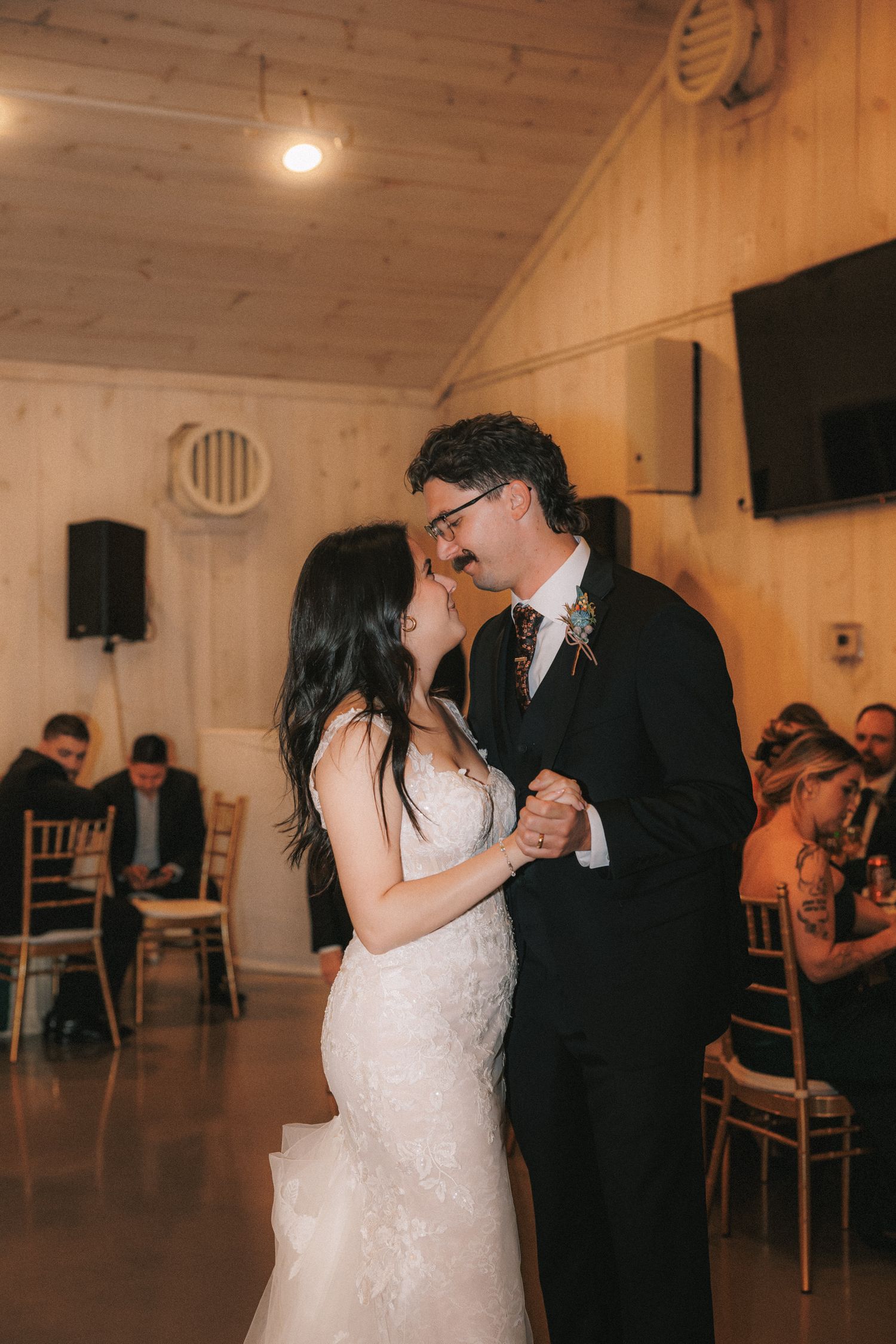 Bride in lace gown and groom in black suit sharing first dance in warmly lit barn venue.