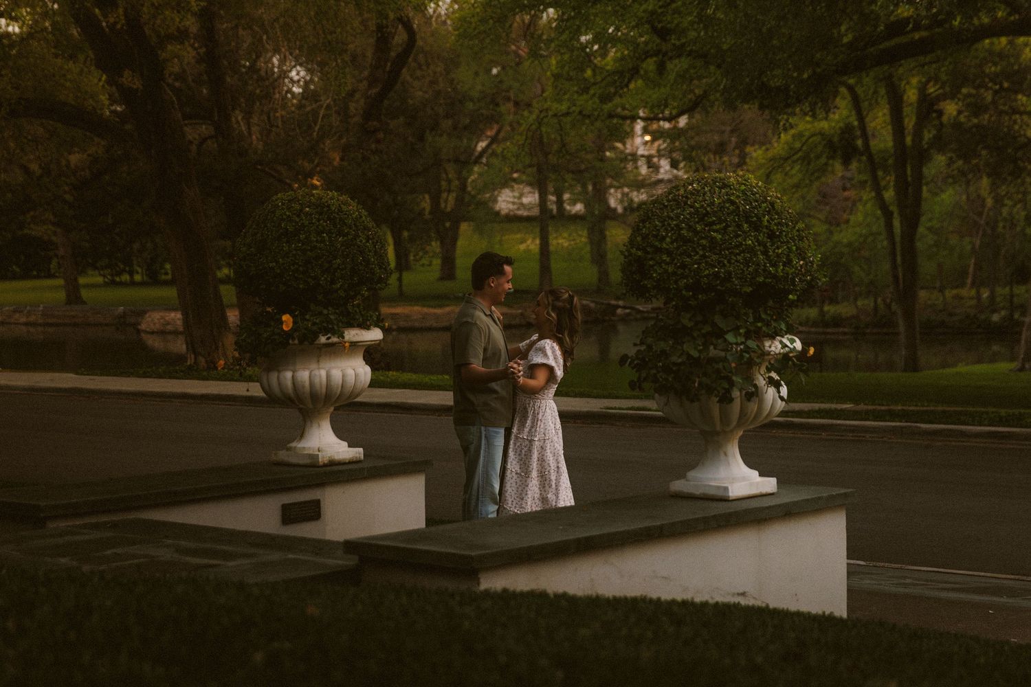 Couple standing together on a garden platform surrounded by lush topiary and white urns at dusk.