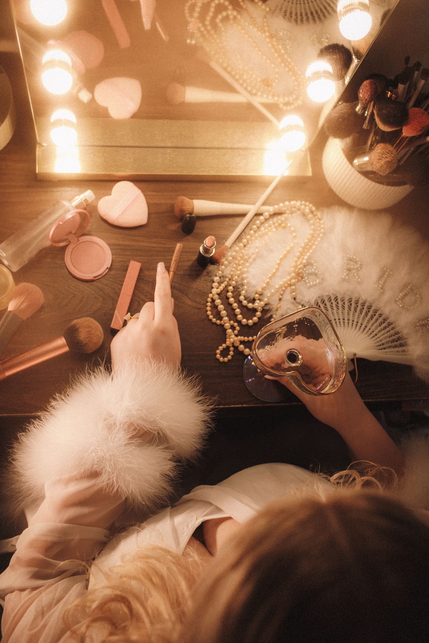 Overhead view of bride at vanity mirror with pearls, fan, perfume, and glowing Hollywood lights.