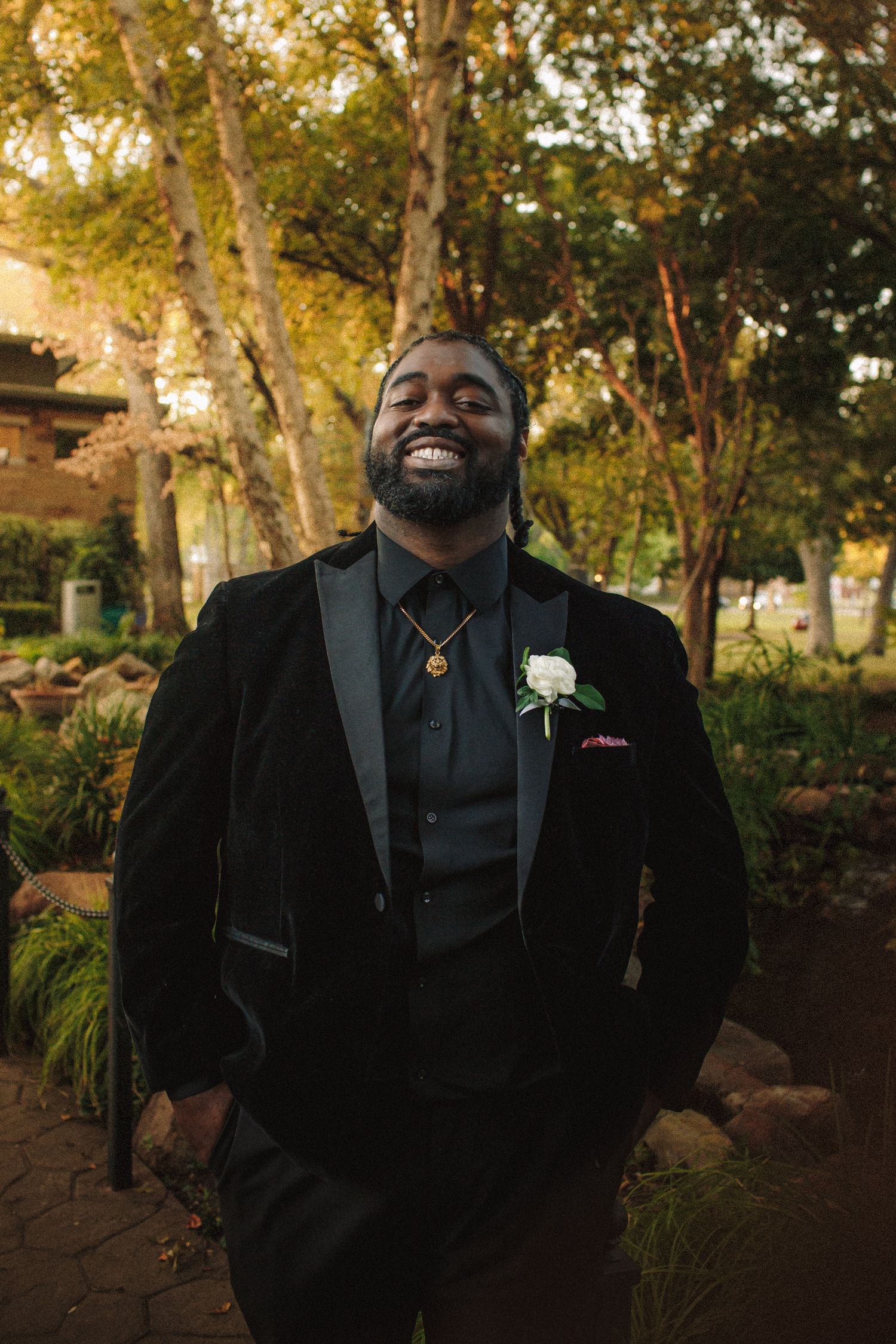 Smiling groom in black velvet tuxedo with white boutonniere poses outdoors in a sunlit garden setting.