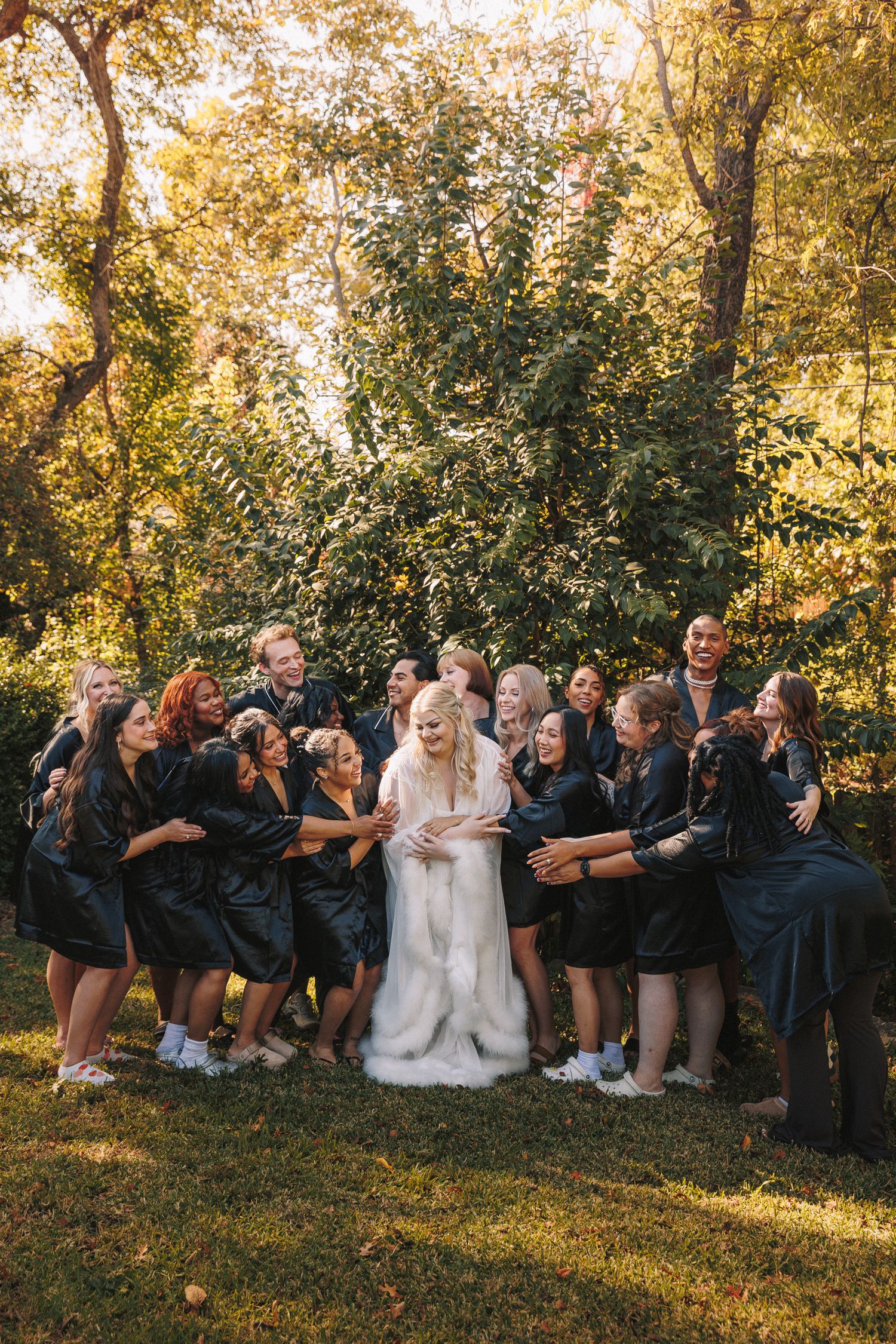 Bride surrounded by joyful bridal party in dark robes sharing a fun group hug in a sunlit garden.