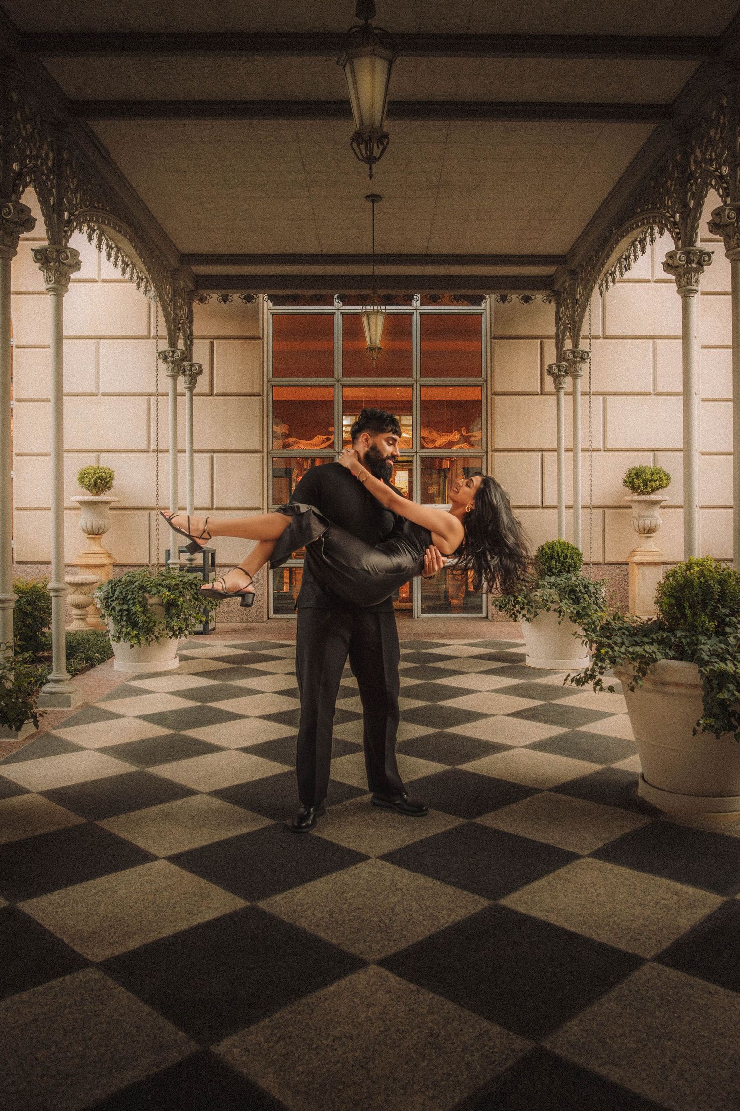 A couple dances gracefully on a checkered floor in an elegant hallway with ornate architecture.
