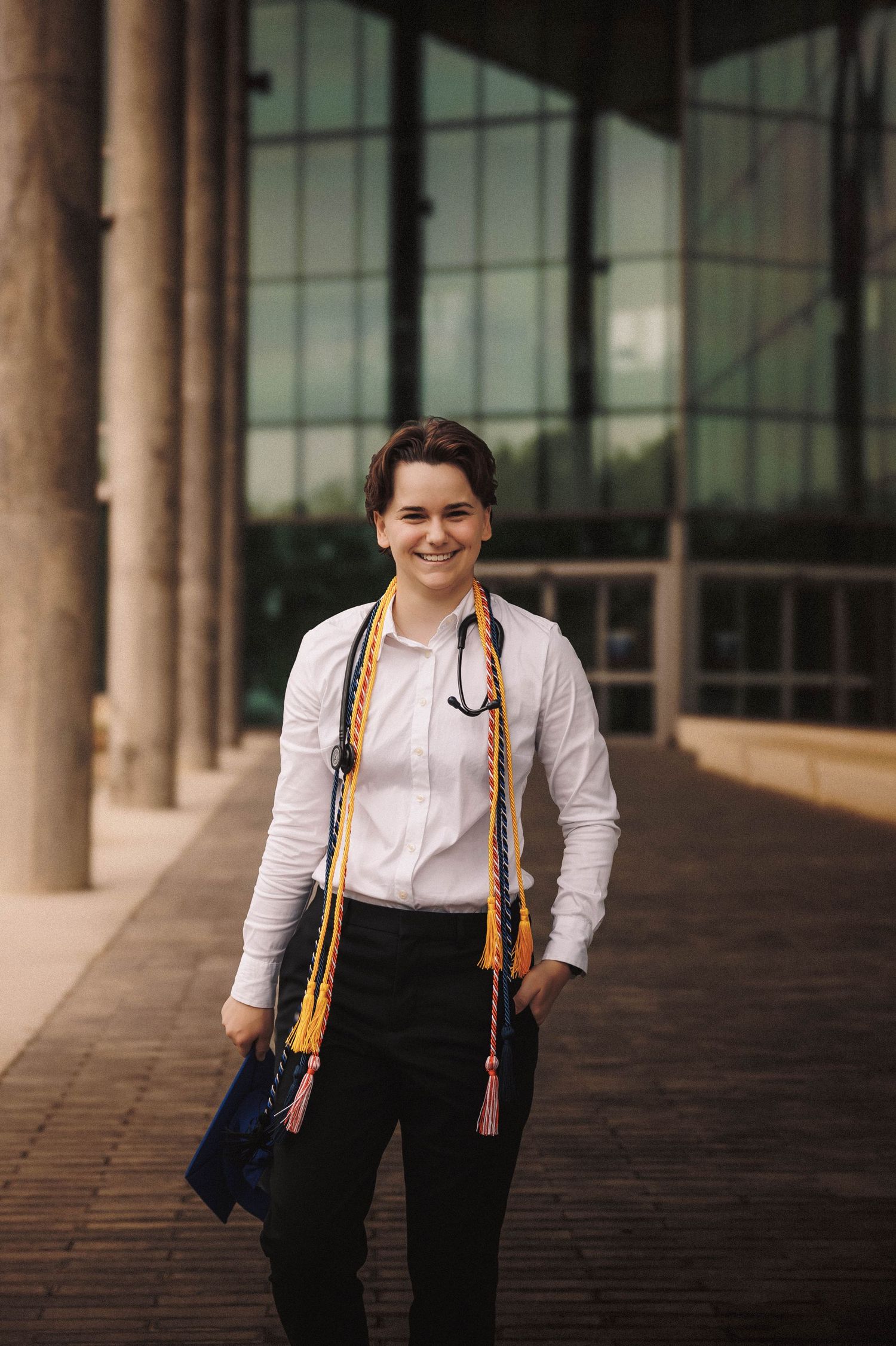 Graduate in formal attire with orange suspenders poses in front of columned building entrance.