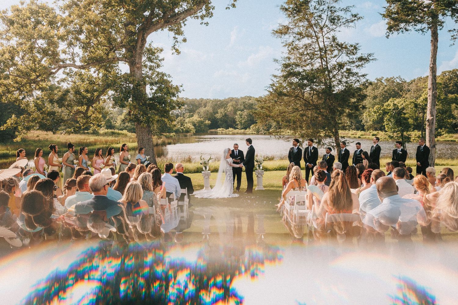 Wedding guests seated in white chairs for an outdoor ceremony by a lake.