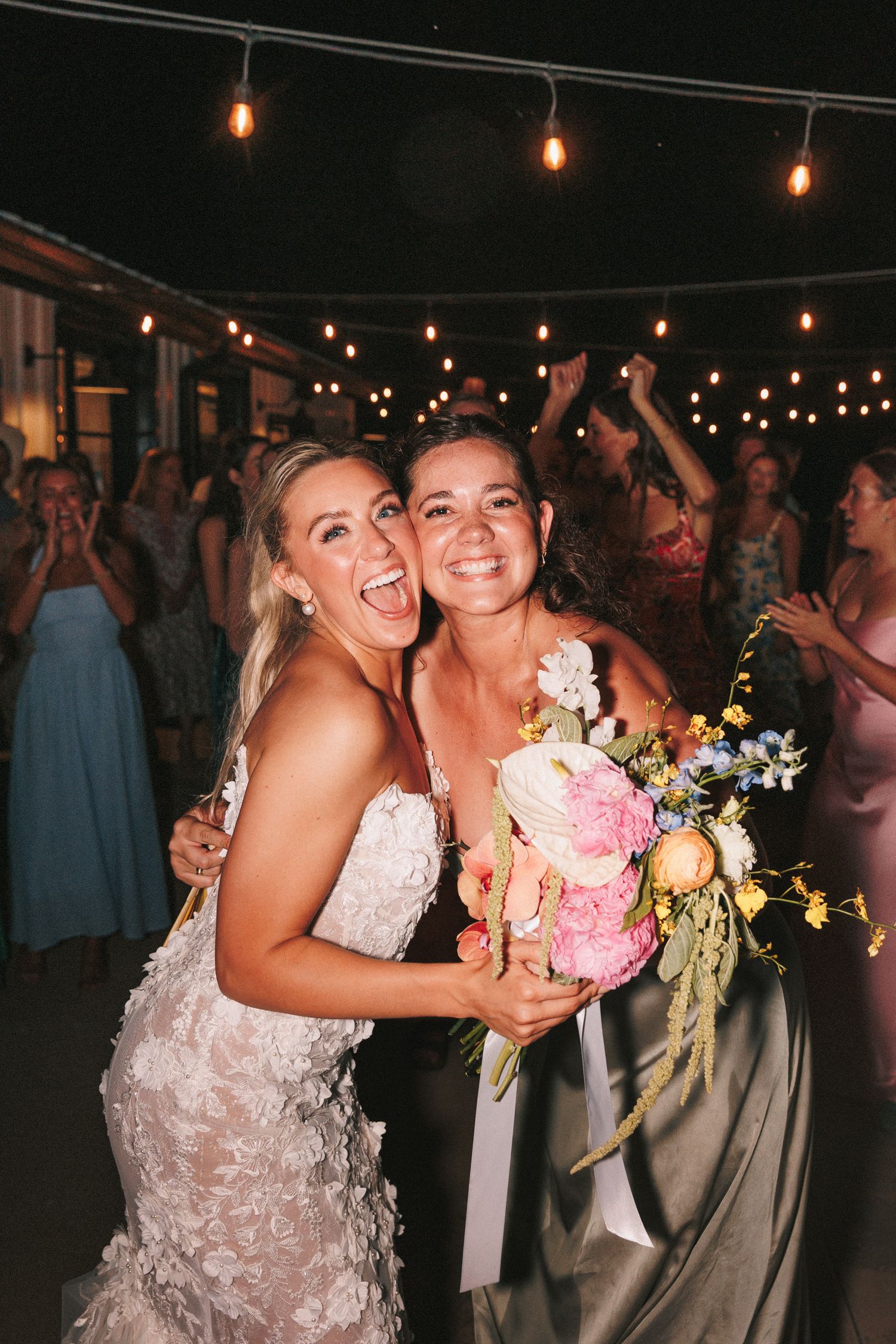 Two people in wedding attire share a joyful moment under string lights while holding a pink floral bouquet at an evening celebration.