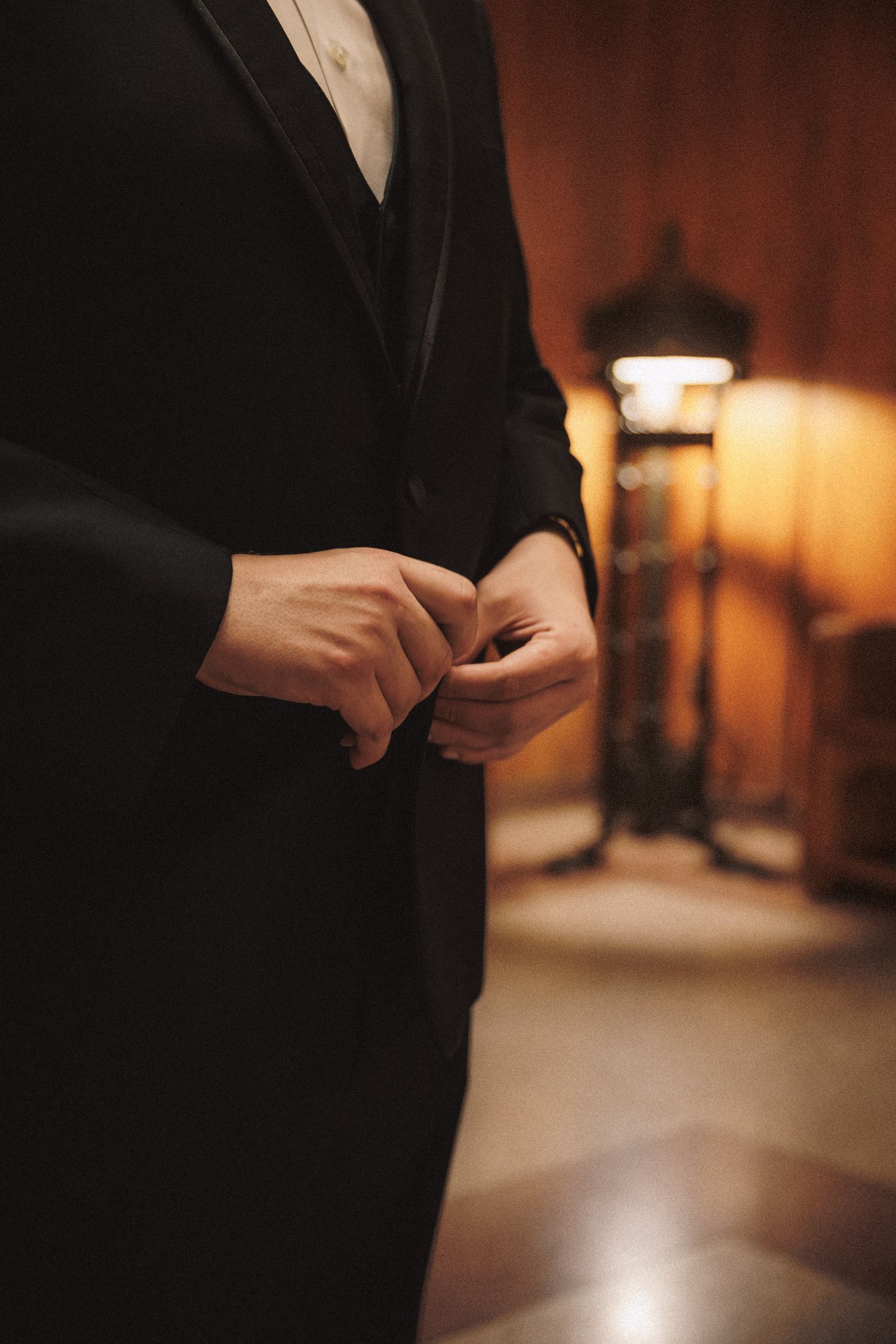 Groom in black tuxedo buttoning jacket before wedding ceremony in warmly lit room.