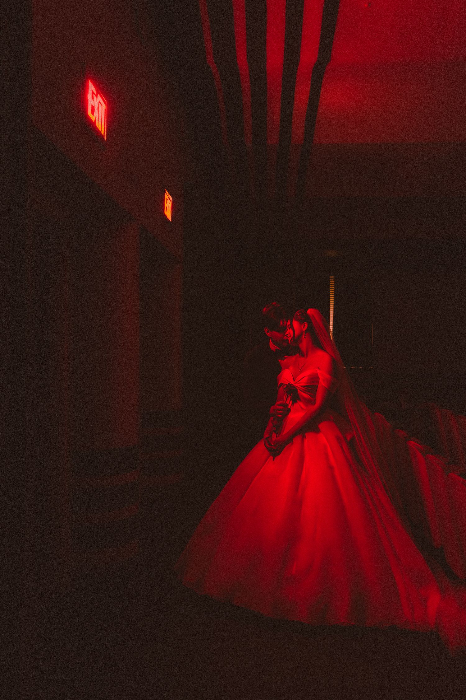 Bride in red ball gown glowing under red light near an exit sign in a dark hallway.