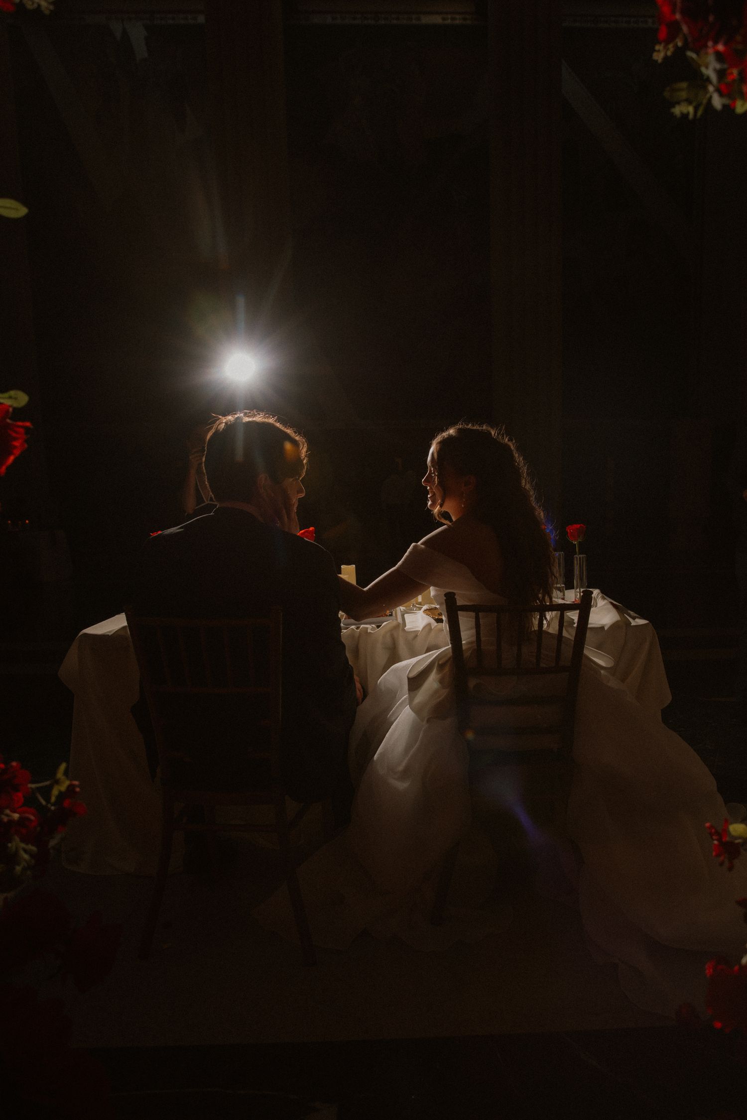 Bride and groom silhouetted at reception table with dramatic spotlight in dark elegant venue.