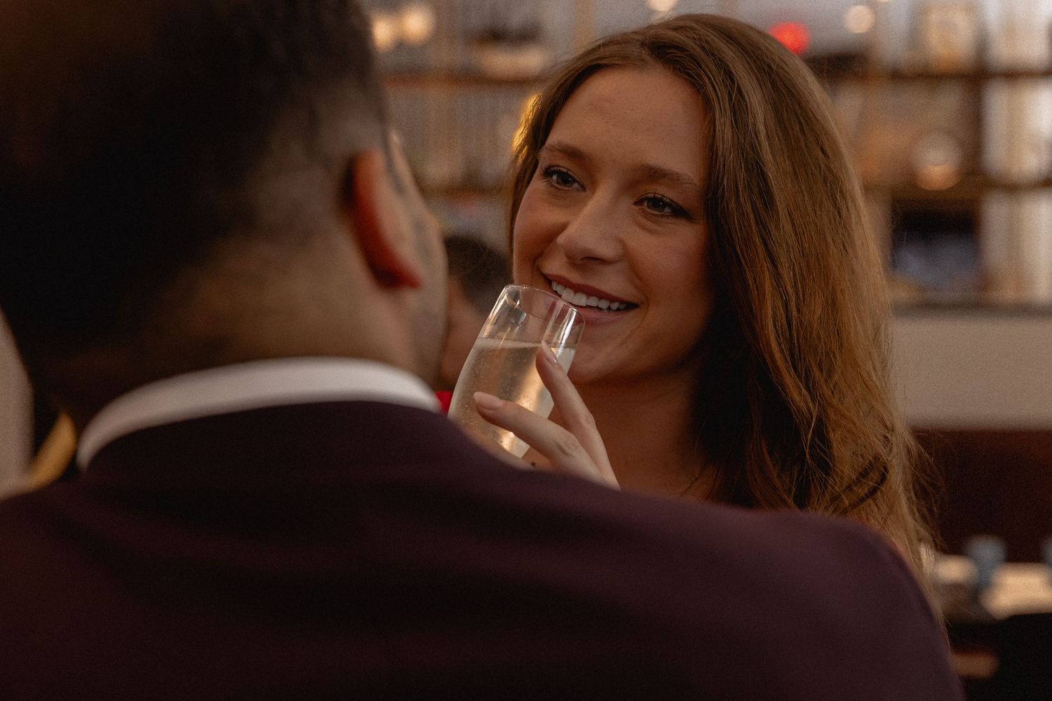 Woman smiling and holding a champagne glass on a romantic date at a dimly lit bar or restaurant.