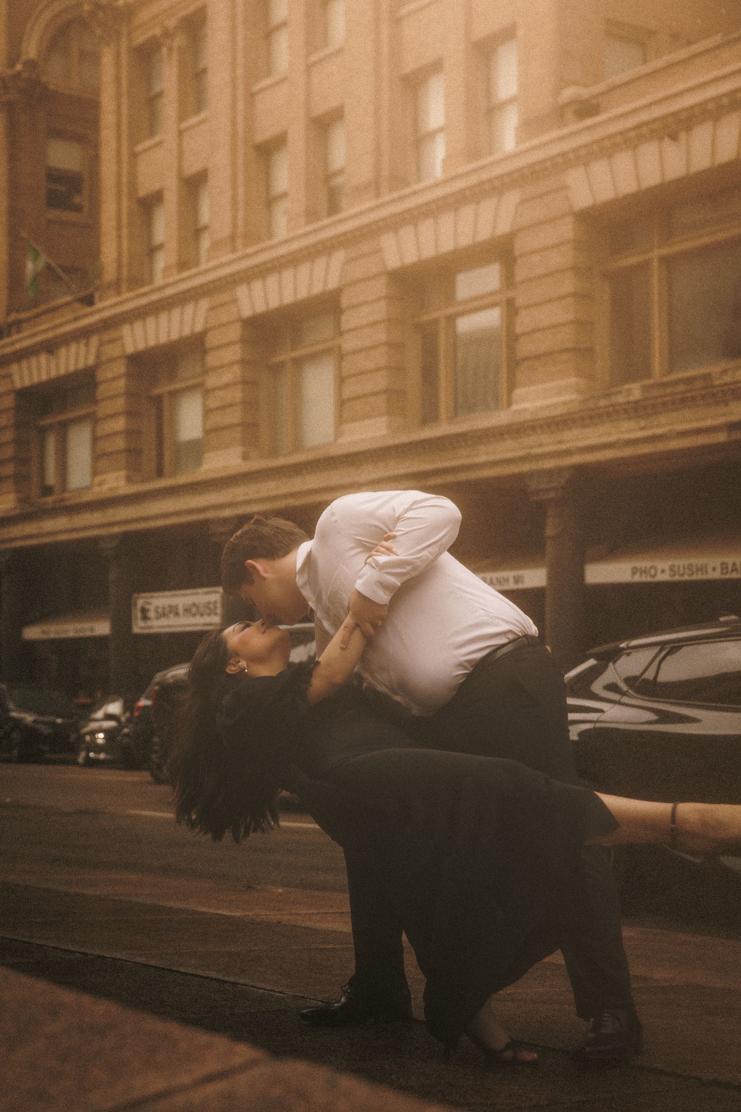 A couple shares a romantic dip kiss on a city street, bathed in warm golden light with historic buildings behind them.
