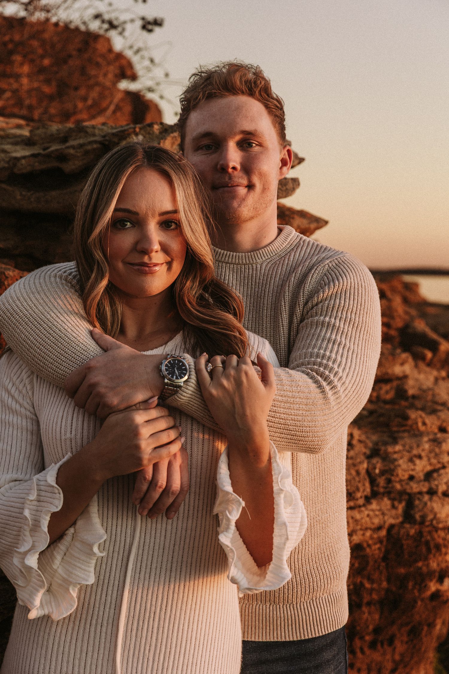 Couple in matching white sweaters embracing on rocky terrain during sunset.