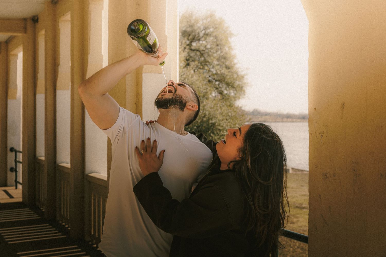 Two people sharing a playful moment on a porch as one drinks from a bottle during sunset.