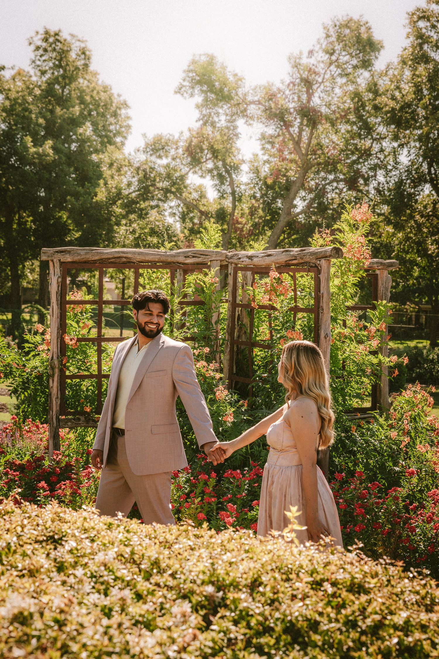 A couple stands in a garden surrounded by blooming white and red flowers under a wooden pergola in golden sunlight.