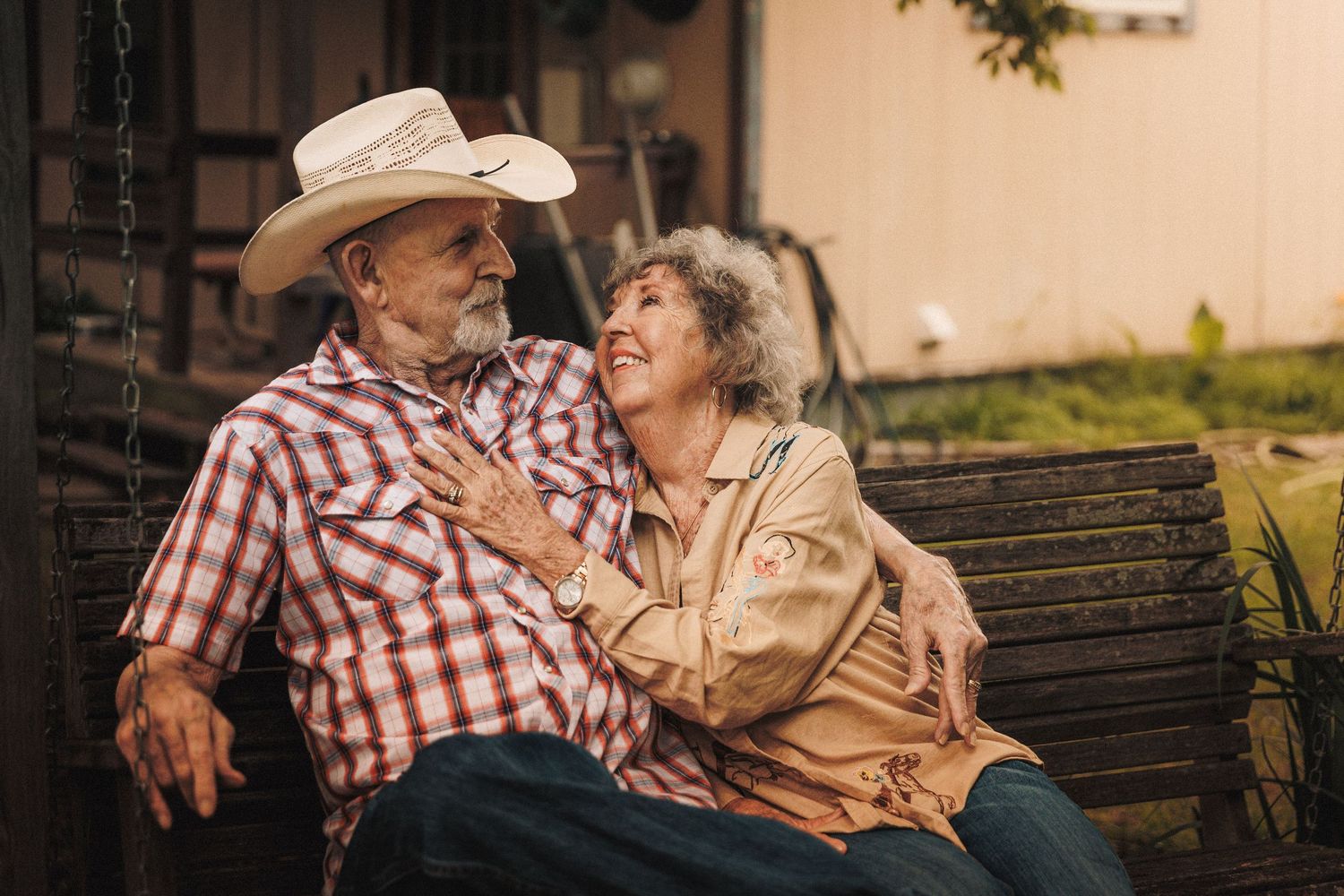 An elderly couple in casual western attire shares a tender moment while sitting together on a bench in a park setting.